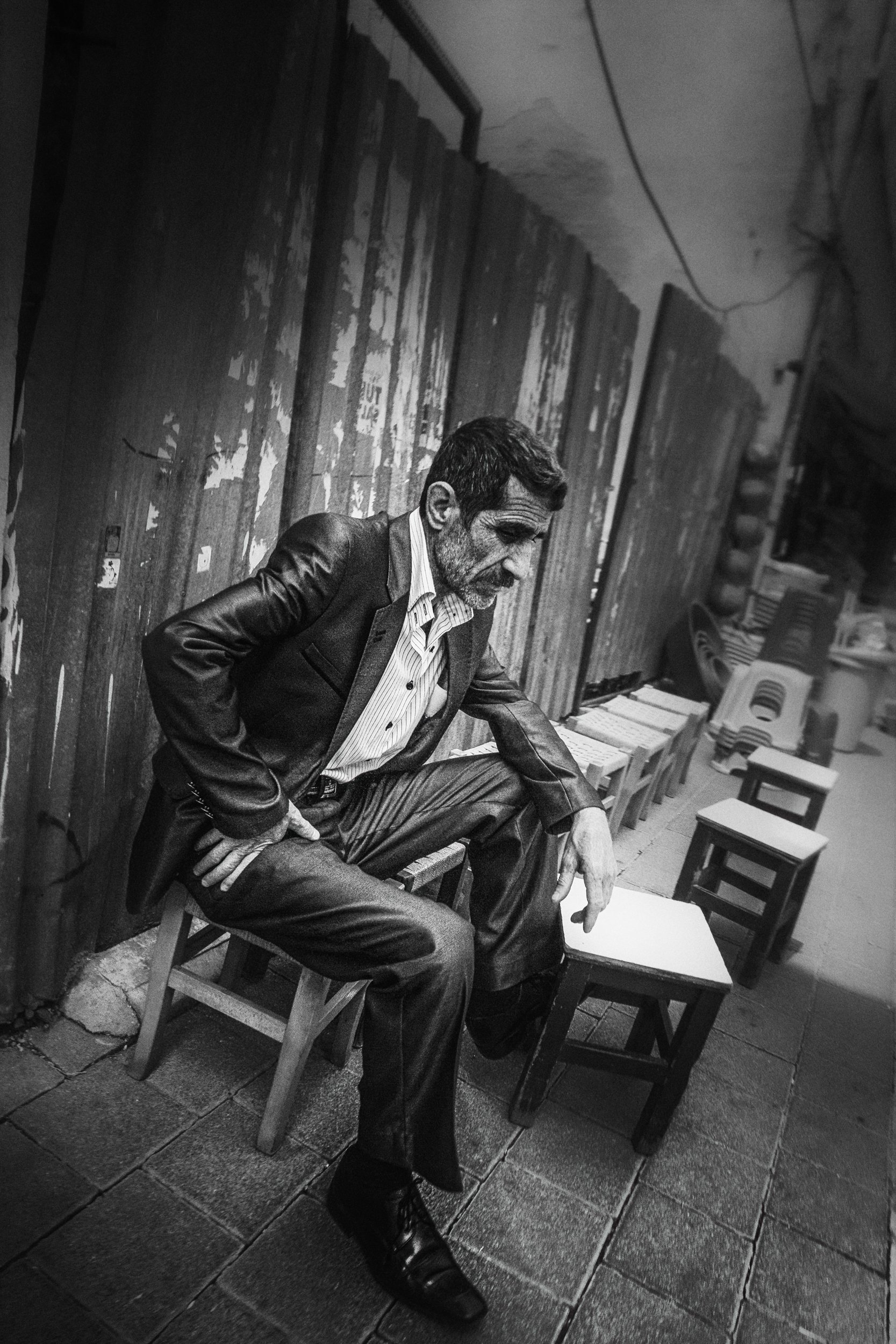 A man wearing a leather jacket, dress shirt, and dress pants, sitting on a small wooden chair on a tiled floor, with multiple small empty chairs lined up beside and behind him, against a rustic wooden wall with peeling paint.