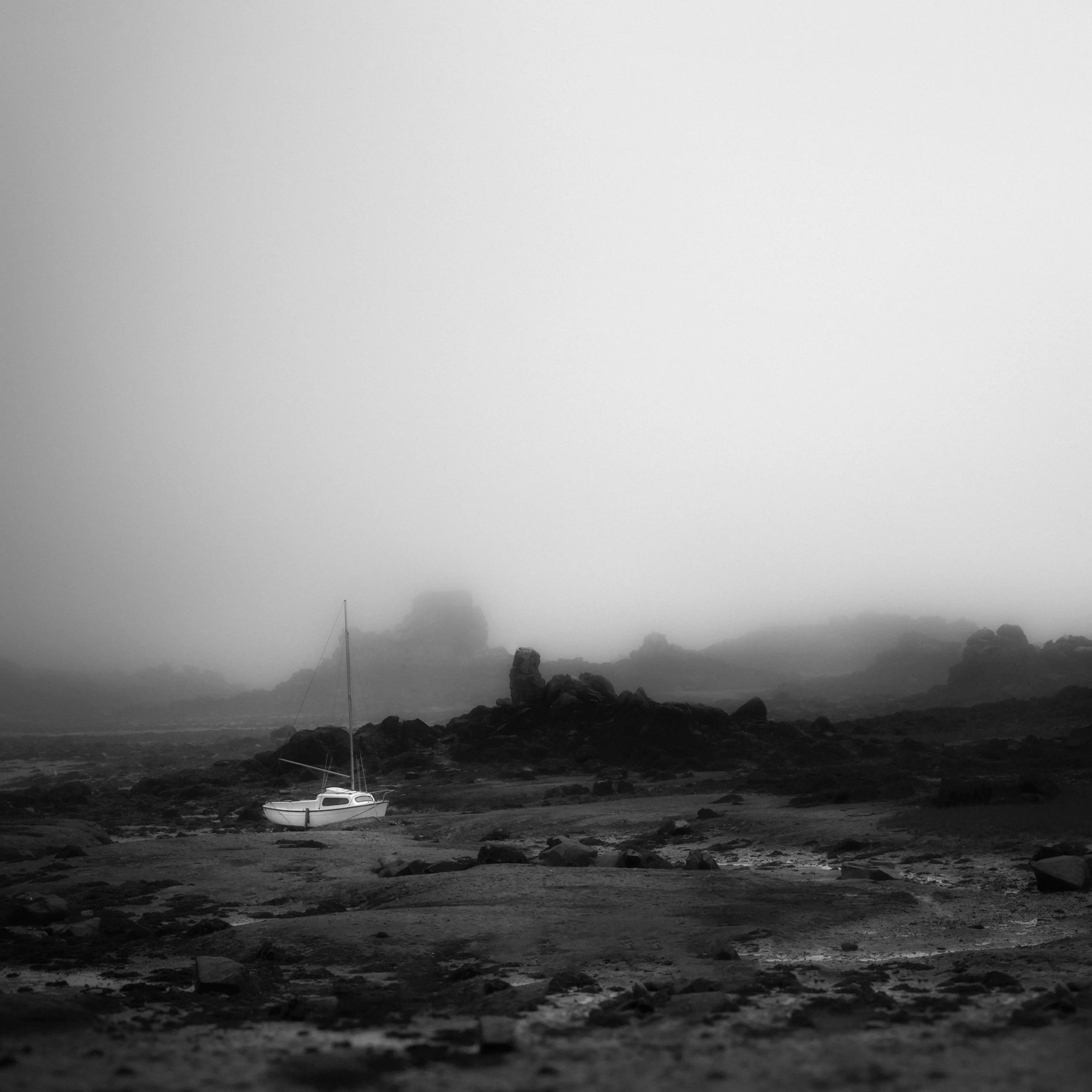 Black and white photo of a lone sailboat grounded on rocky shoreline with overcast sky and foggy background.
