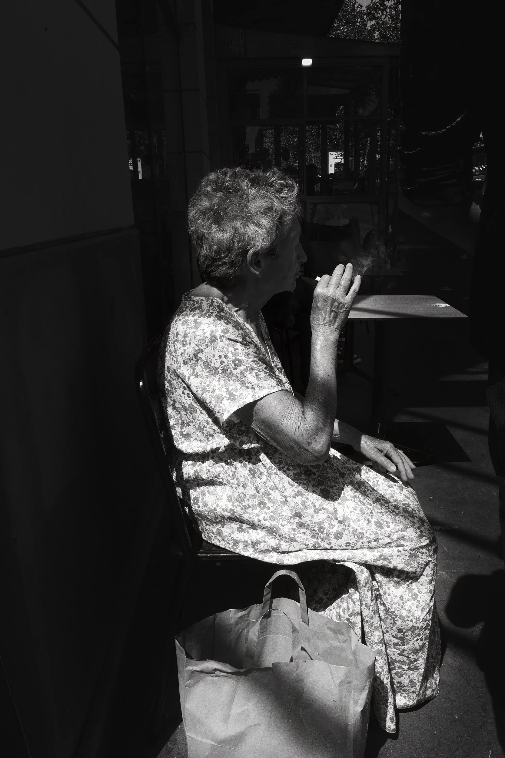 A black and white photograph of an elderly woman with short, curly hair sitting at a table outside a café or restaurant. She is wearing a floral dress and is in the process of smoking a cigarette, with smoke rising from it. There is a paper bag on th