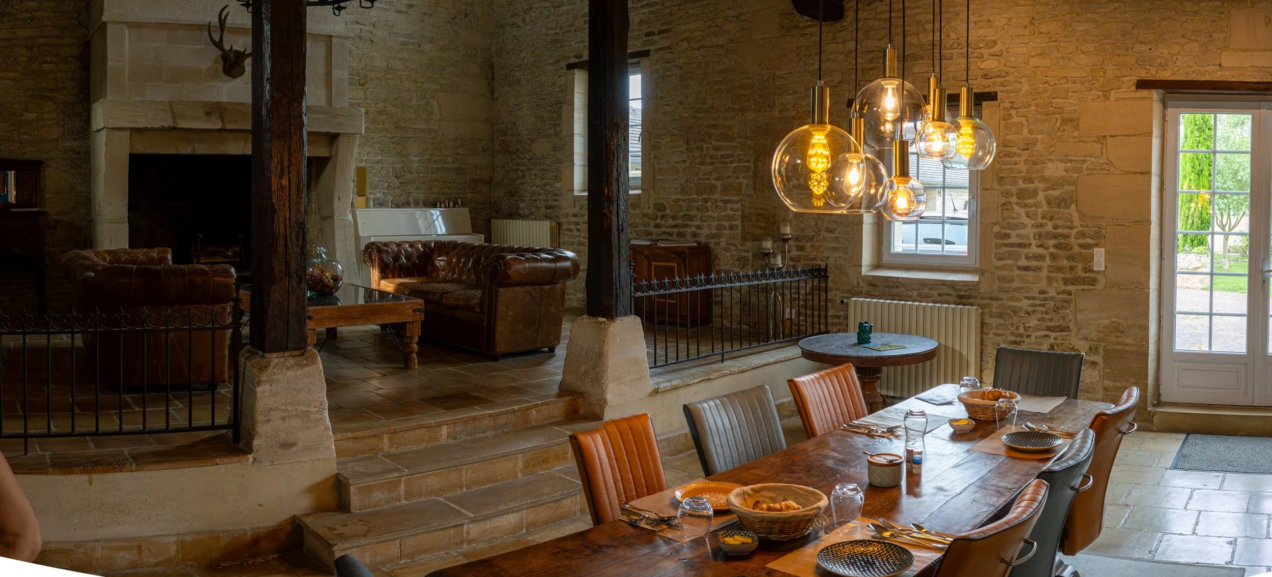 Interior of a rustic living and dining area with exposed brick walls, wooden beams, a stone fireplace with a mounted deer head, leather chairs, a wooden dining table set with bowls and glasses, a cluster of hanging glass pendant lights, and windows w