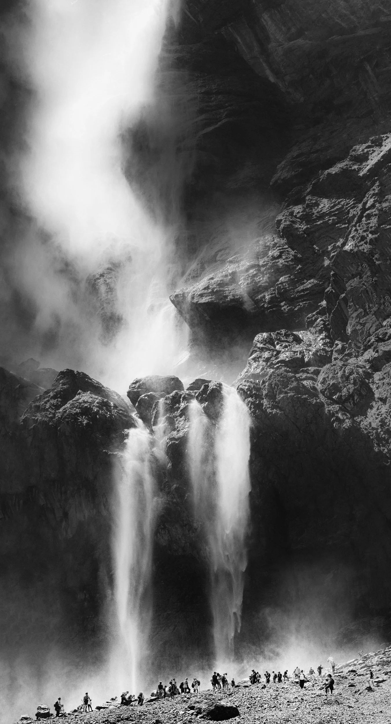 Black and white photo of a large waterfall with a mist at the bottom, and a group of people standing on rocky terrain nearby.