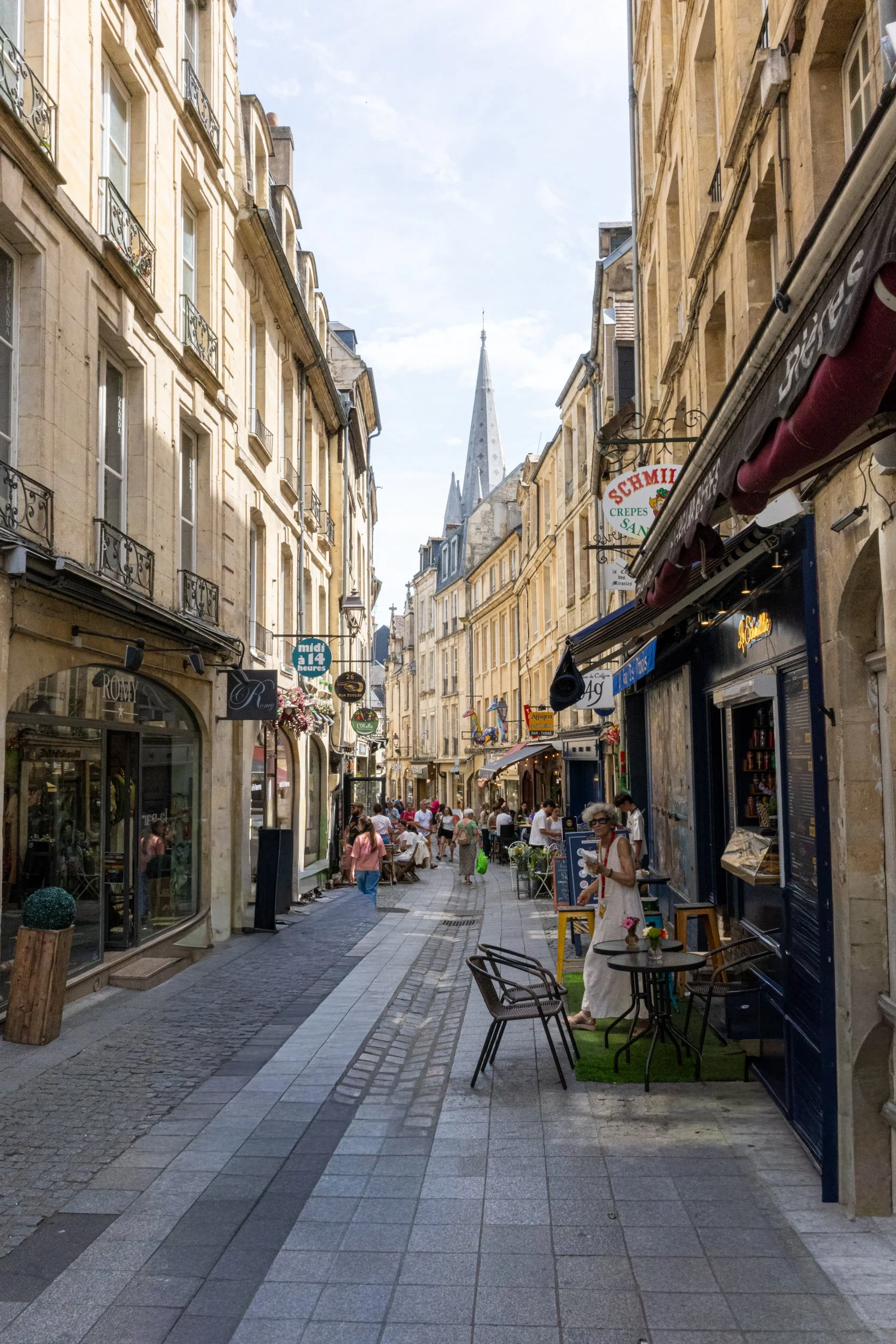 A busy pedestrian street lined with shops and cafes, with people walking and sitting outdoors; historic buildings with ornate balconies; church steeples visible in the background; clear sky with a few clouds.