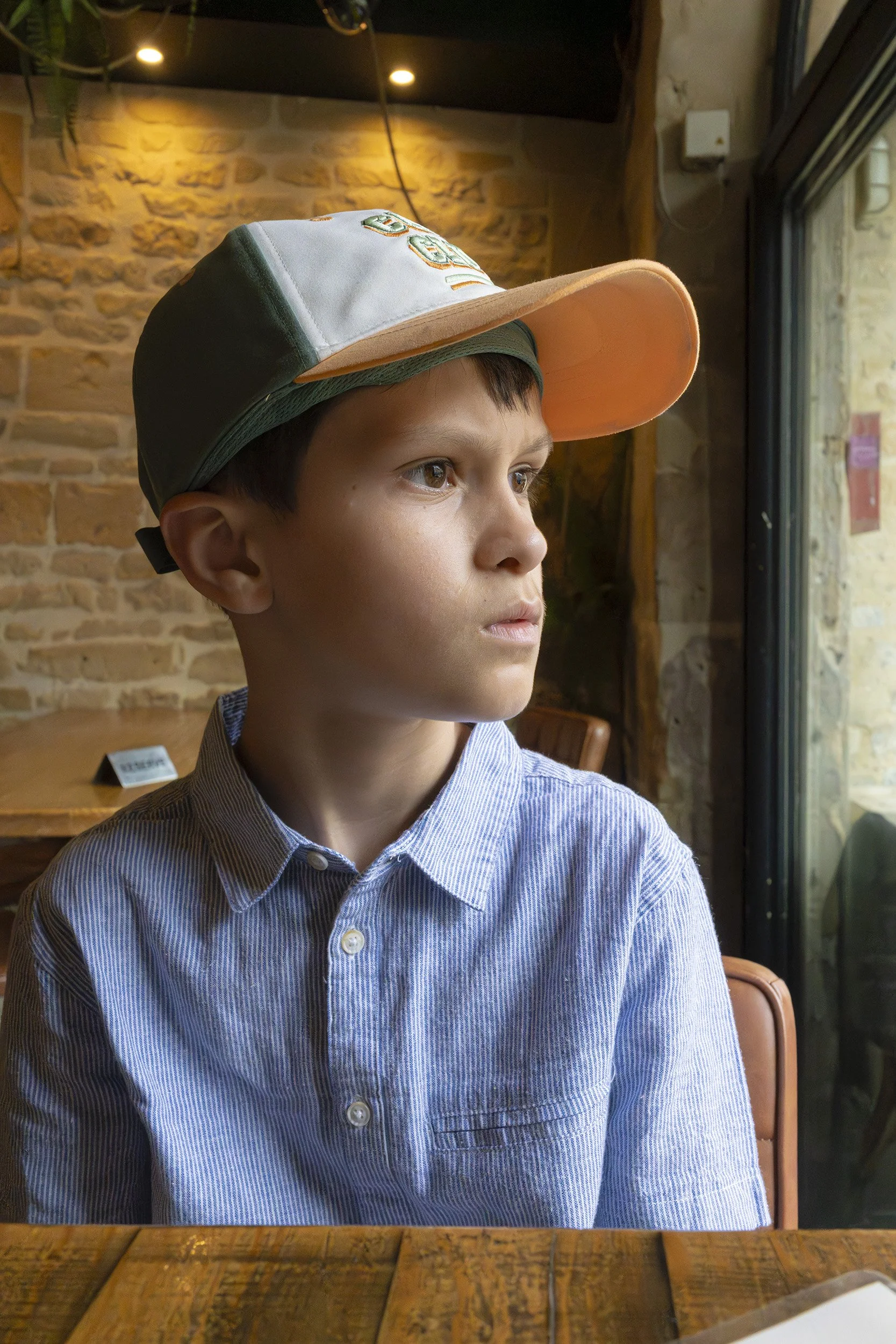 A young boy with short dark hair and light brown skin, wearing a blue and white striped button-up shirt and a colorful trucker hat, sitting in a restaurant or café. The background features a brick wall with warm lighting and a window to his right.