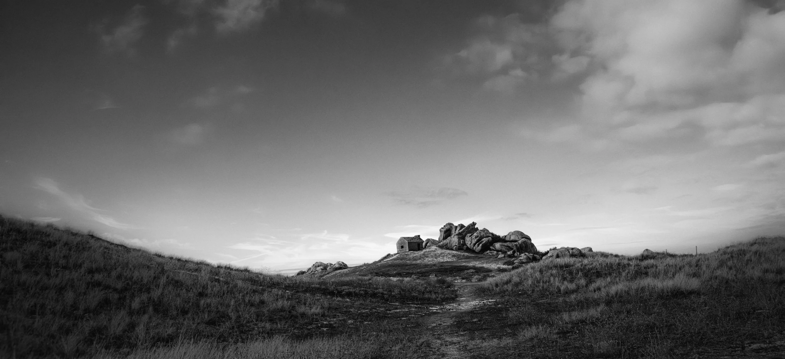 A small stone house surrounded by large rocks on a hill, with grassy fields on either side, under a mostly cloudy sky.
