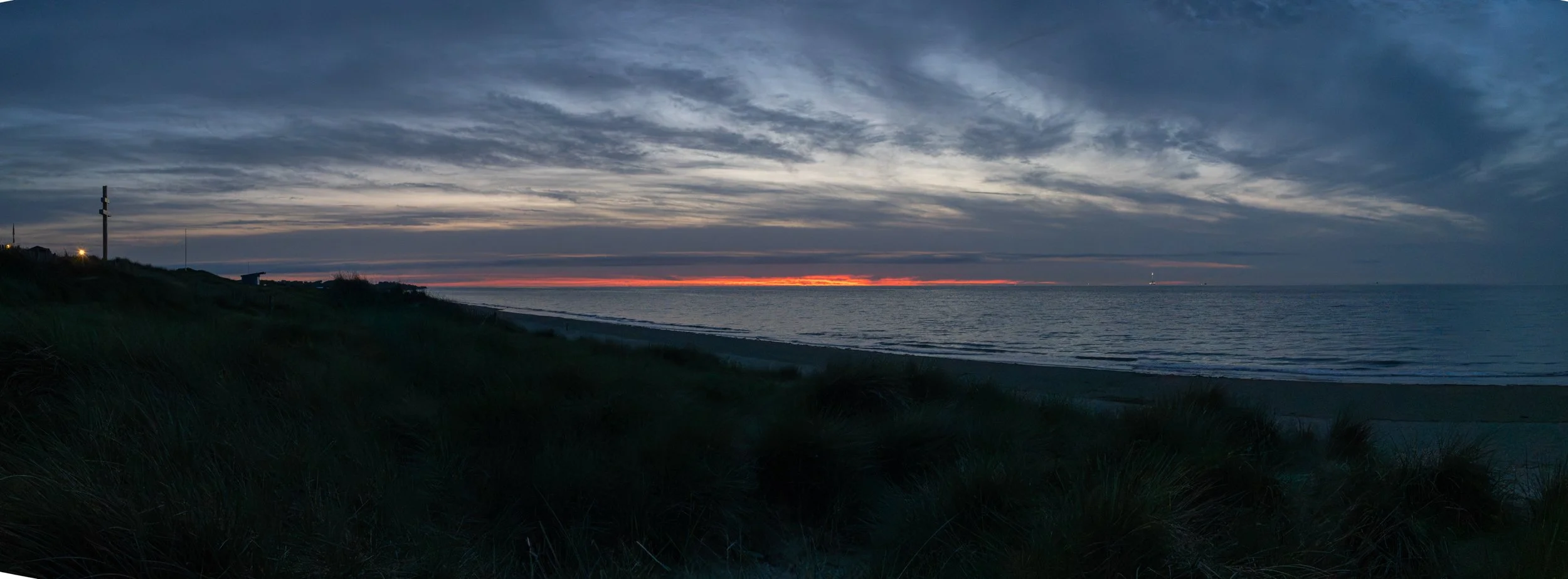 A scenic view of a sunset over the ocean with a partly cloudy sky and a dark shoreline with grass in the foreground.