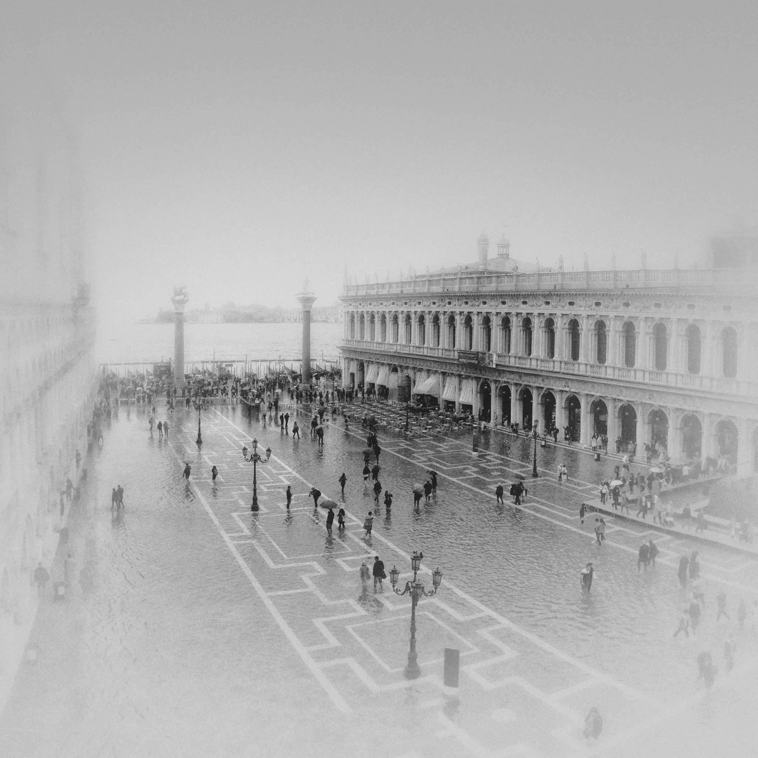 A foggy cityscape with a large historic building, several lampposts, and people walking in a plaza.