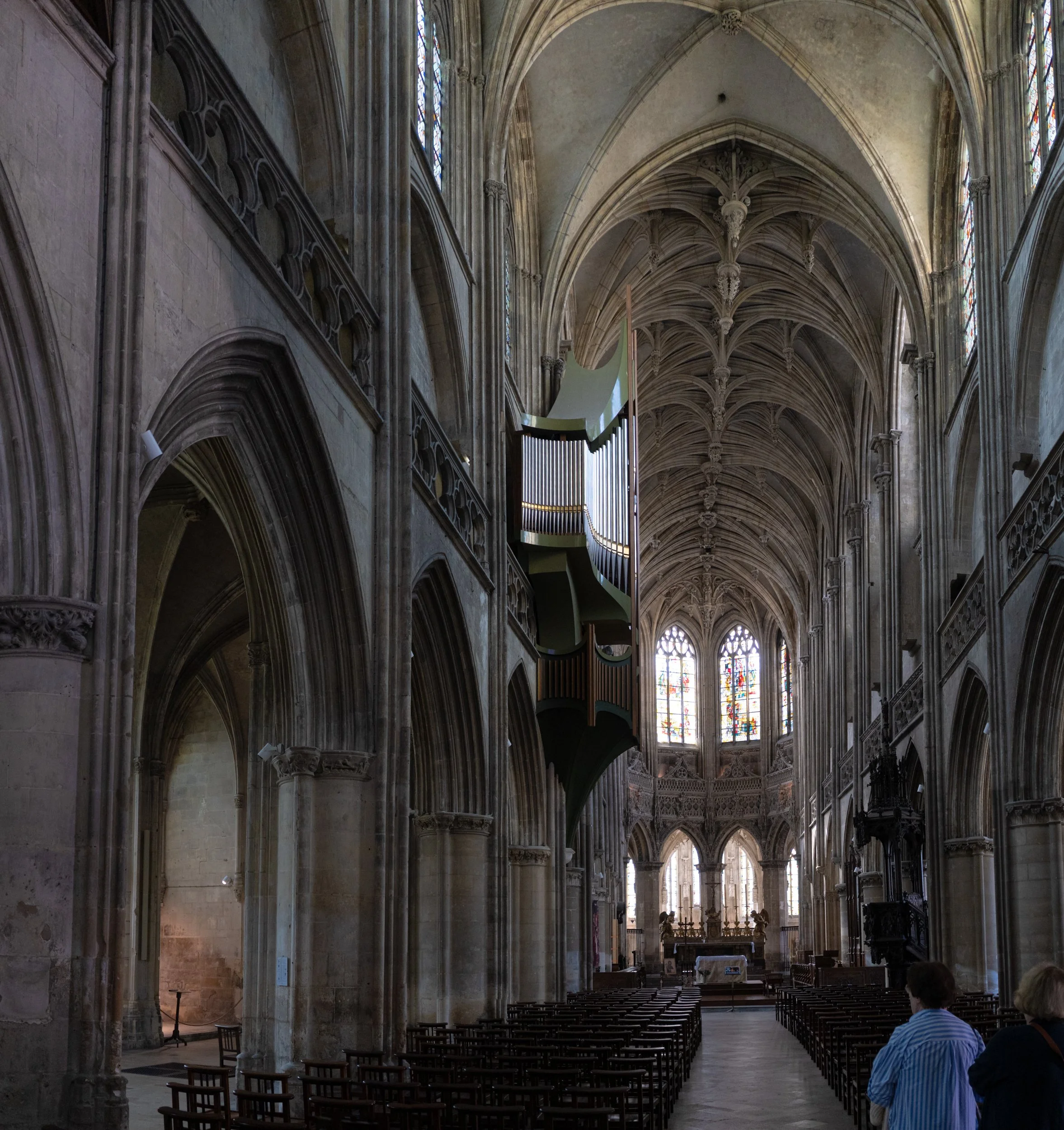 Interior of a historic church with high vaulted ceilings, stained glass windows, stone arches, and rows of wooden chairs. Some people are seated, facing the altar area.