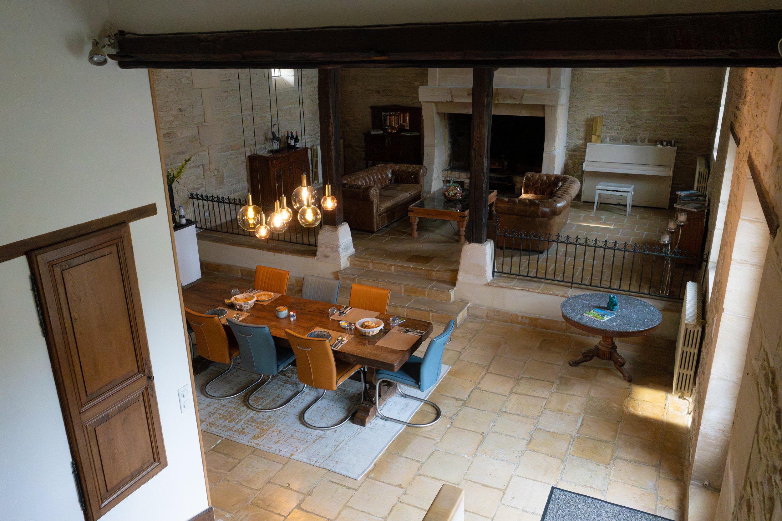 A rustic dining and sitting area in a house with stone walls, leather sofas, a white piano, and a chandelier with exposed bulbs.