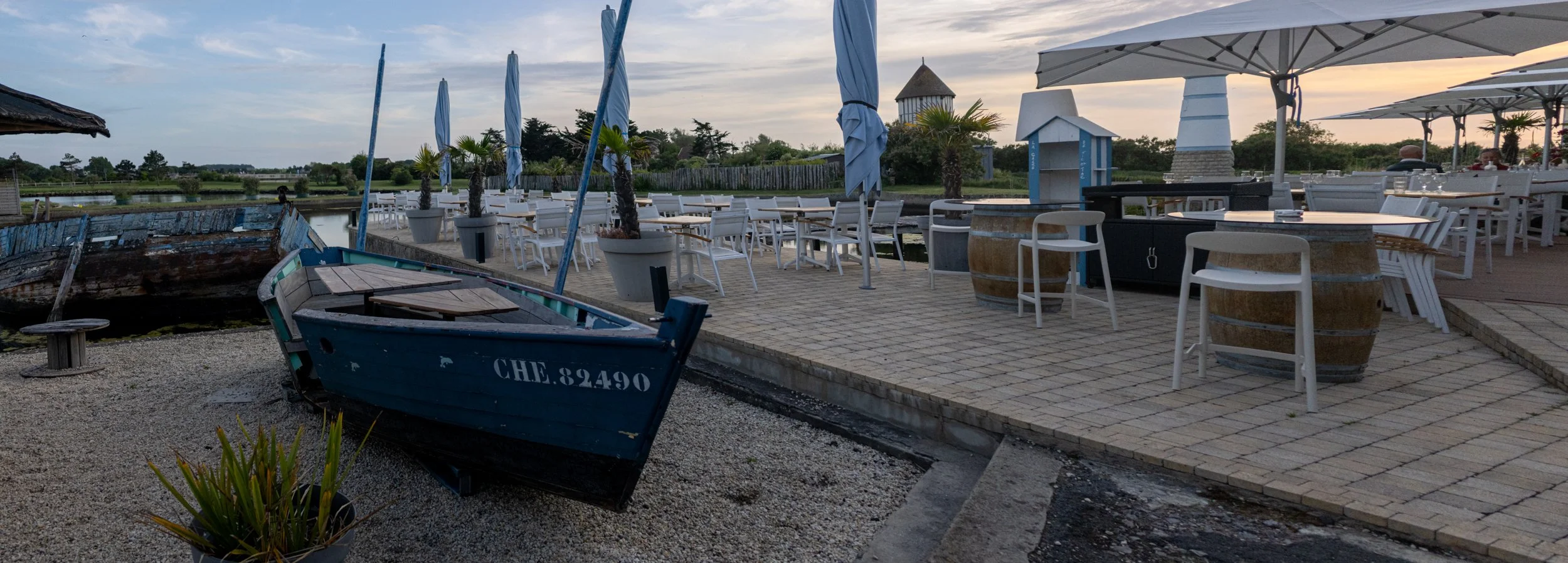 Outdoor dining area on a patio near water with several white chairs, tables, blue umbrellas, potted palm trees, and a small boat in the foreground at dusk.