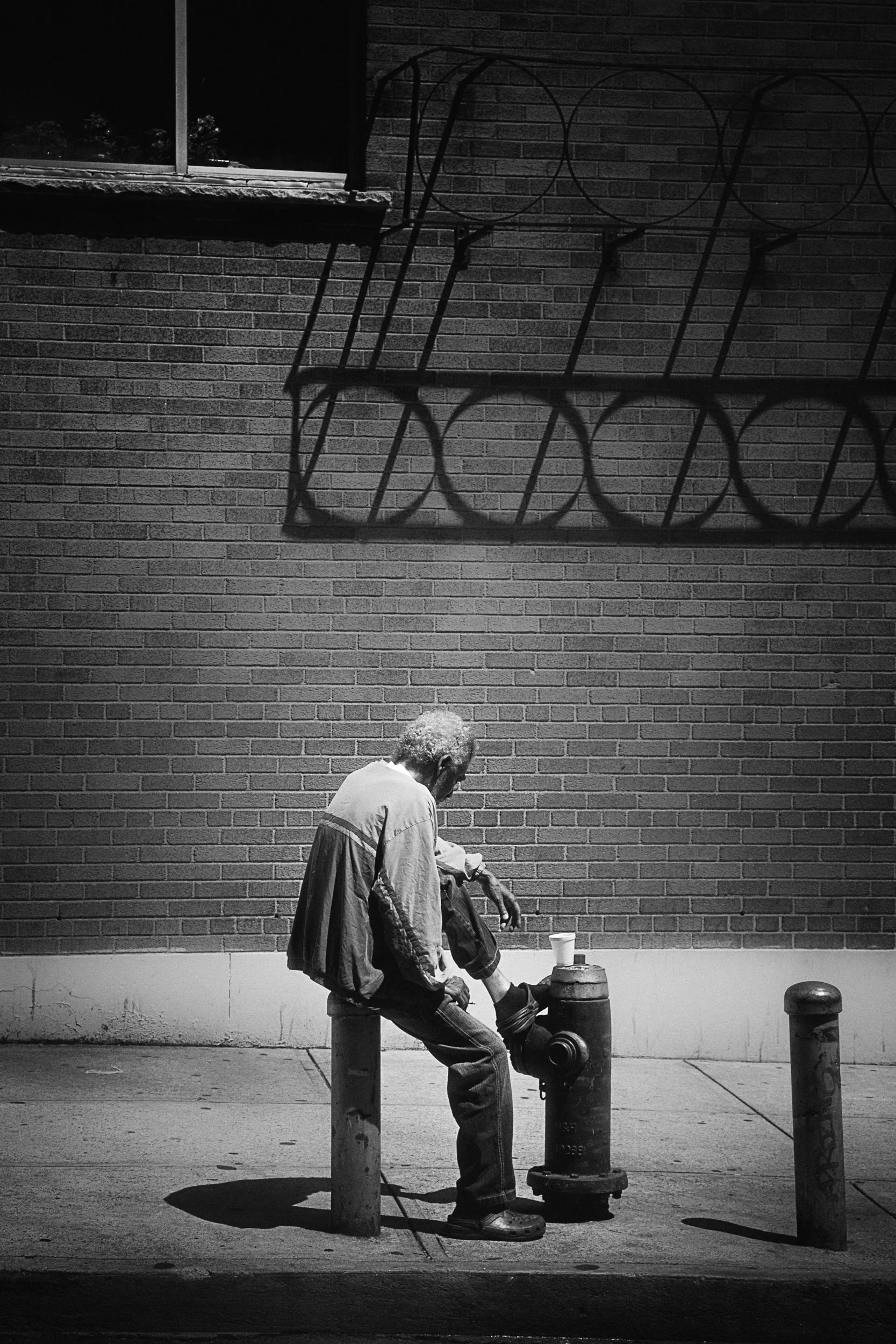 A man with curly hair dressed in a jacket and jeans is sitting on a post on the sidewalk near fire hydrants. The image is in black and white and features a brick wall with a window and metal wire frames casting shadows.