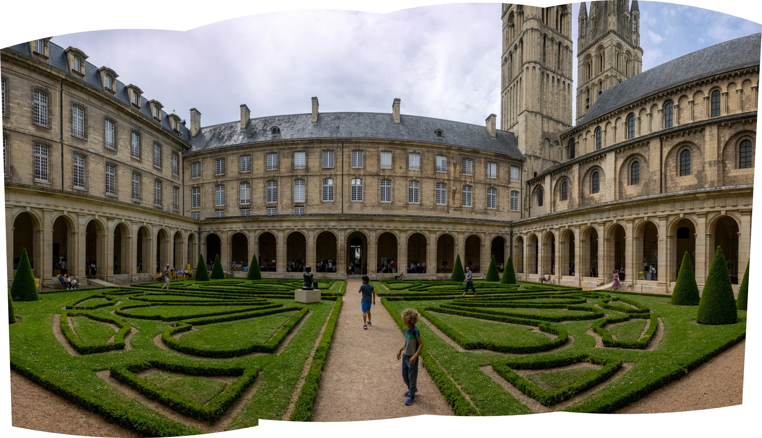 A historic stone building with tall towers and clock faces, surrounded by a manicured garden with spiraled hedges and a gravel pathway, where children are walking and sitting.