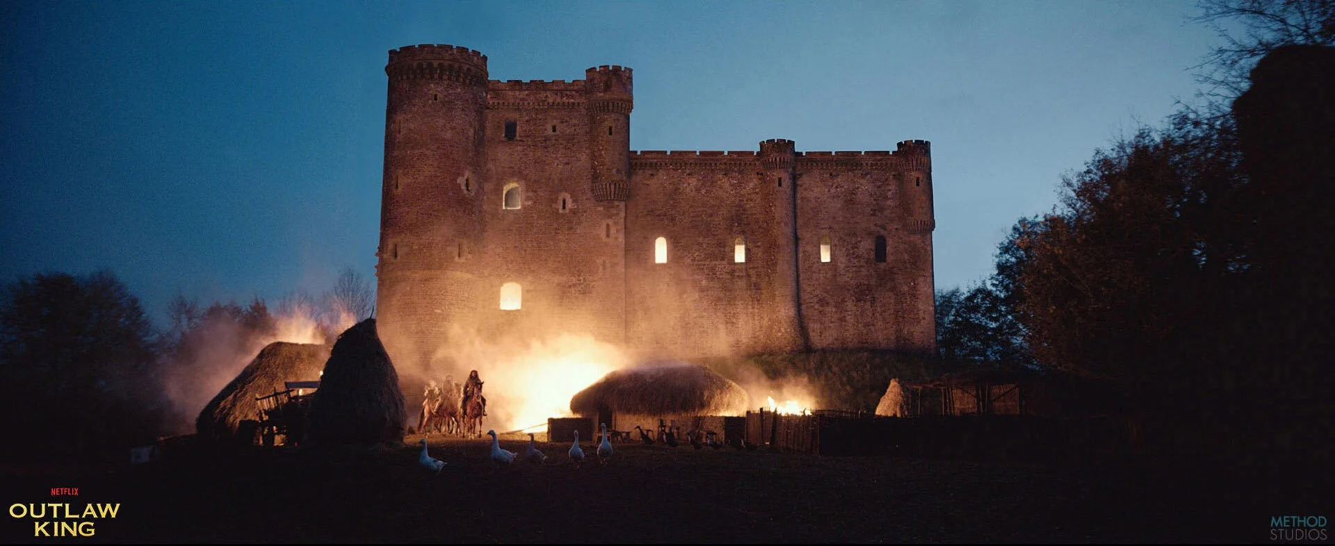 A castle illuminated at night, with flames and smoke rising from the ground in front, and a few horses and white geese nearby. The scene appears to be from a movie or show, with the Netflix Outlaw King logo in the bottom left corner and Method Studio