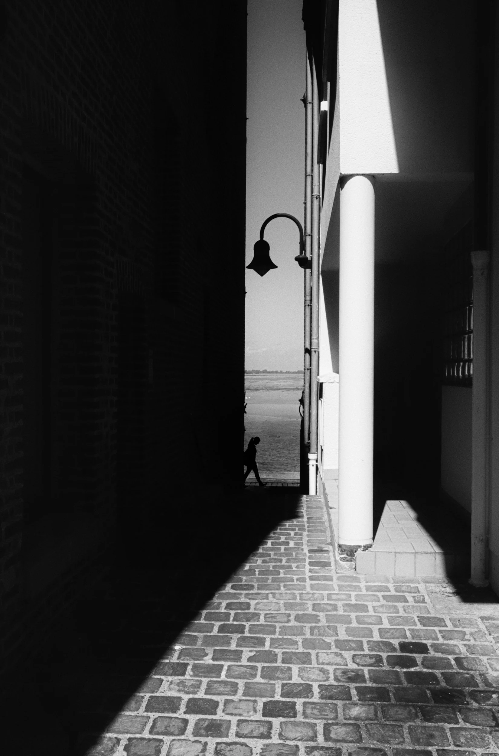 Black and white photo of a narrow alleyway with a brick paved floor, flanked by tall buildings creating shadows and darkness on the left side, with a person walking near the water in the distance outside.