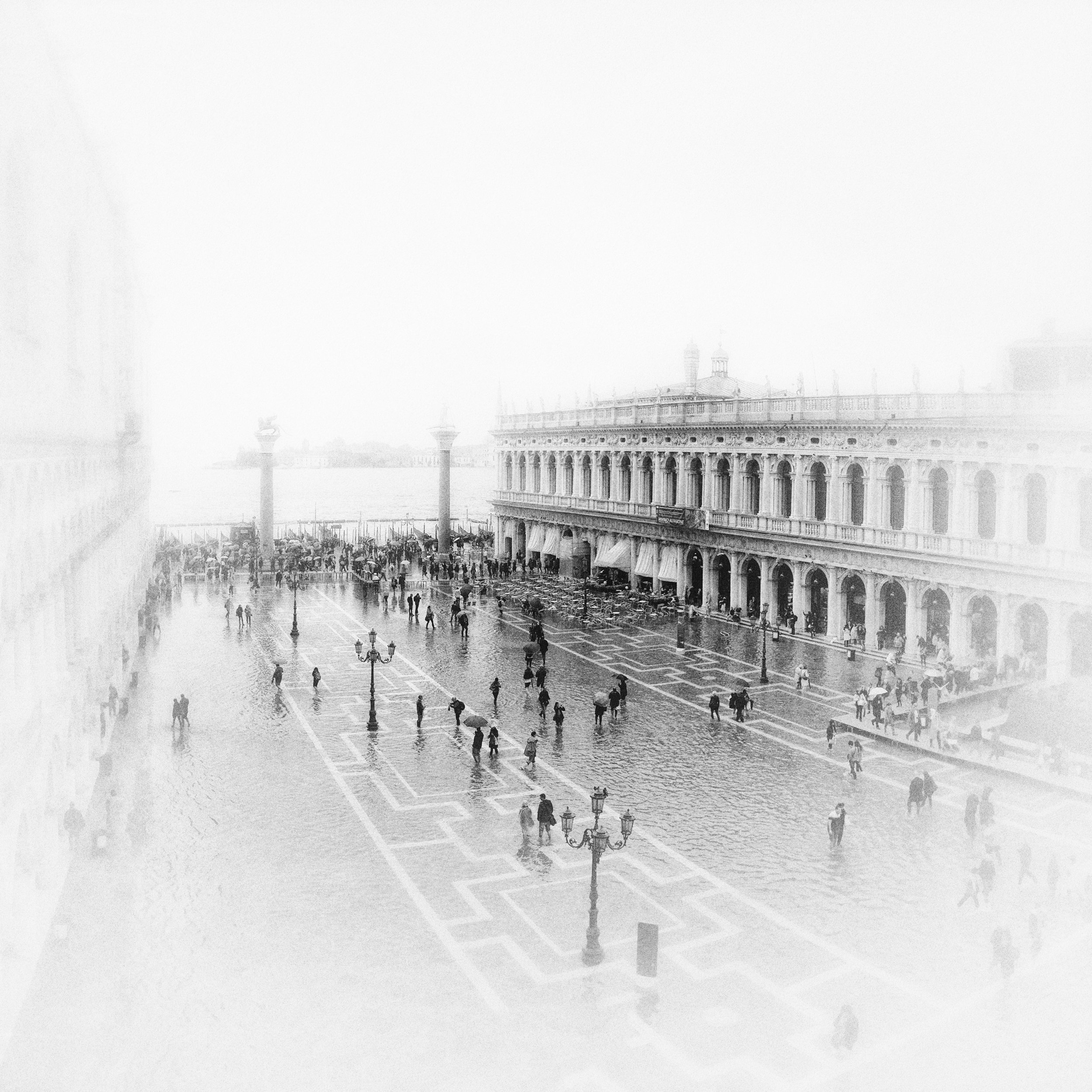 A crowded outdoor plaza with a large building featuring arches and columns, people walking, and street lamps, possibly during a rainy day.