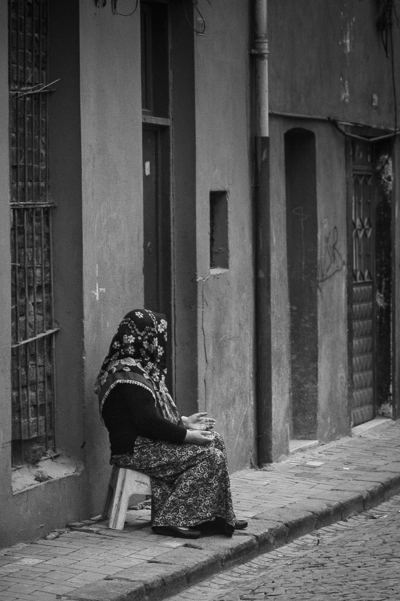 A woman seated on a small stool on a sidewalk, wearing traditional clothing and a headscarf, in front of a building with barred windows and a closed gate.