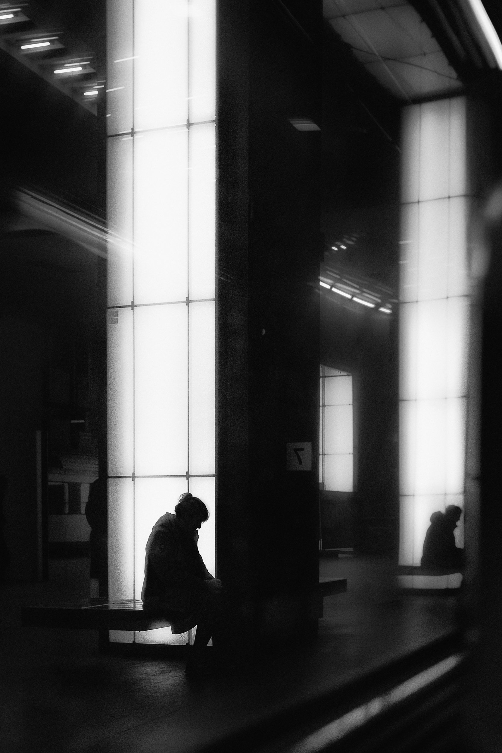 Silhouettes of people sitting on benches in a modern, illuminated indoor space with large backlit panels and reflective surfaces.