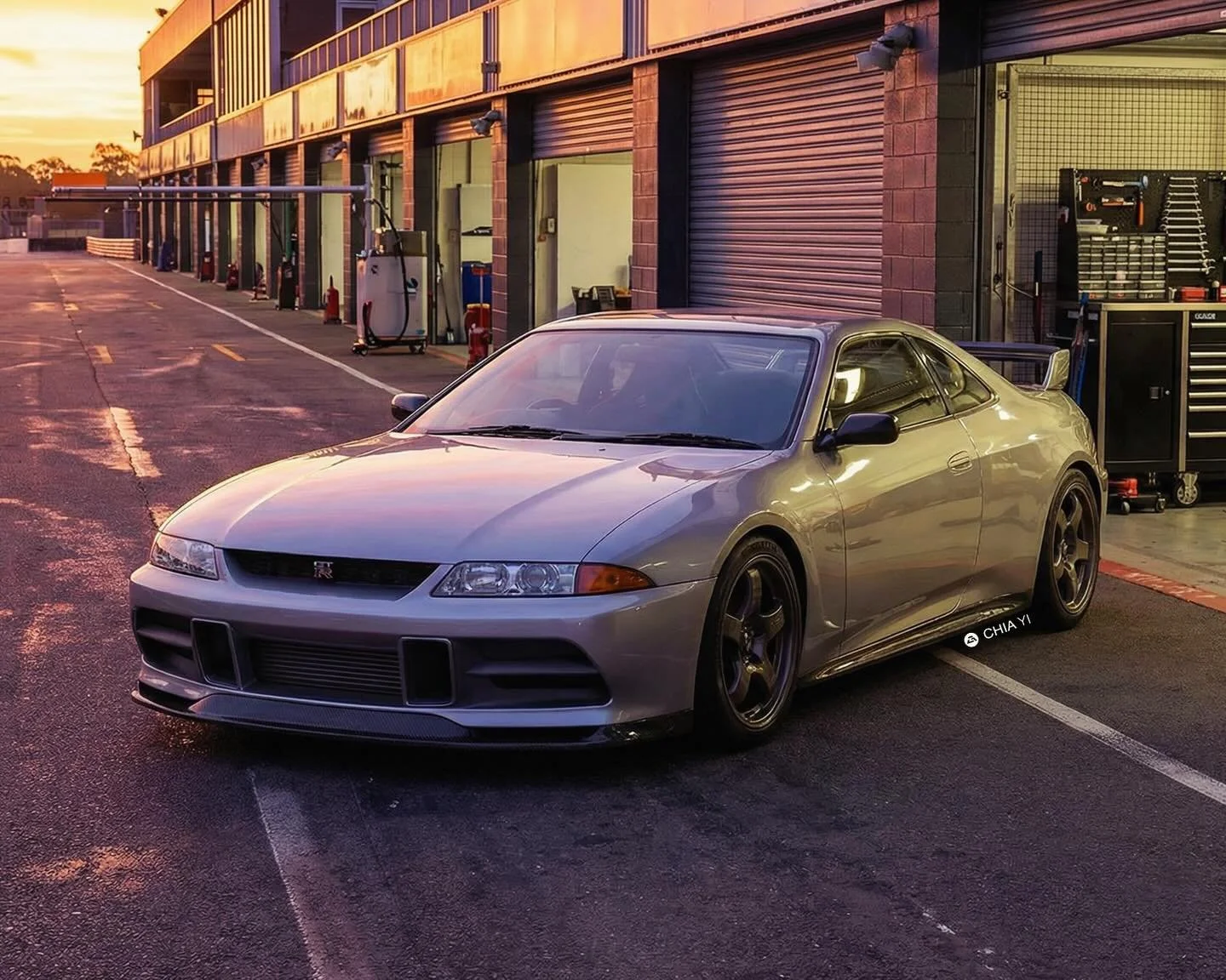 This concept rendering places a modernized 1989 Nissan Skyline GT-R R32 right at home in a sunlit race track pit lane. The sleek silver bodywork reflects the warm evening glow, highlighting subtly widened fenders and refined aerodynamics. Up front, t