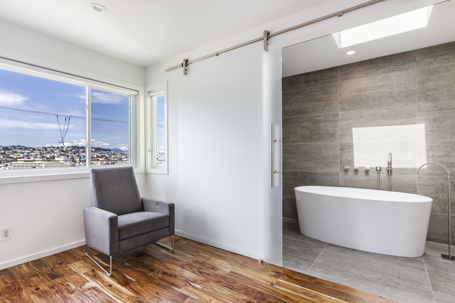bathroom with glass barn door and grey tile and white tub and brushed nickel fixtures.jpg