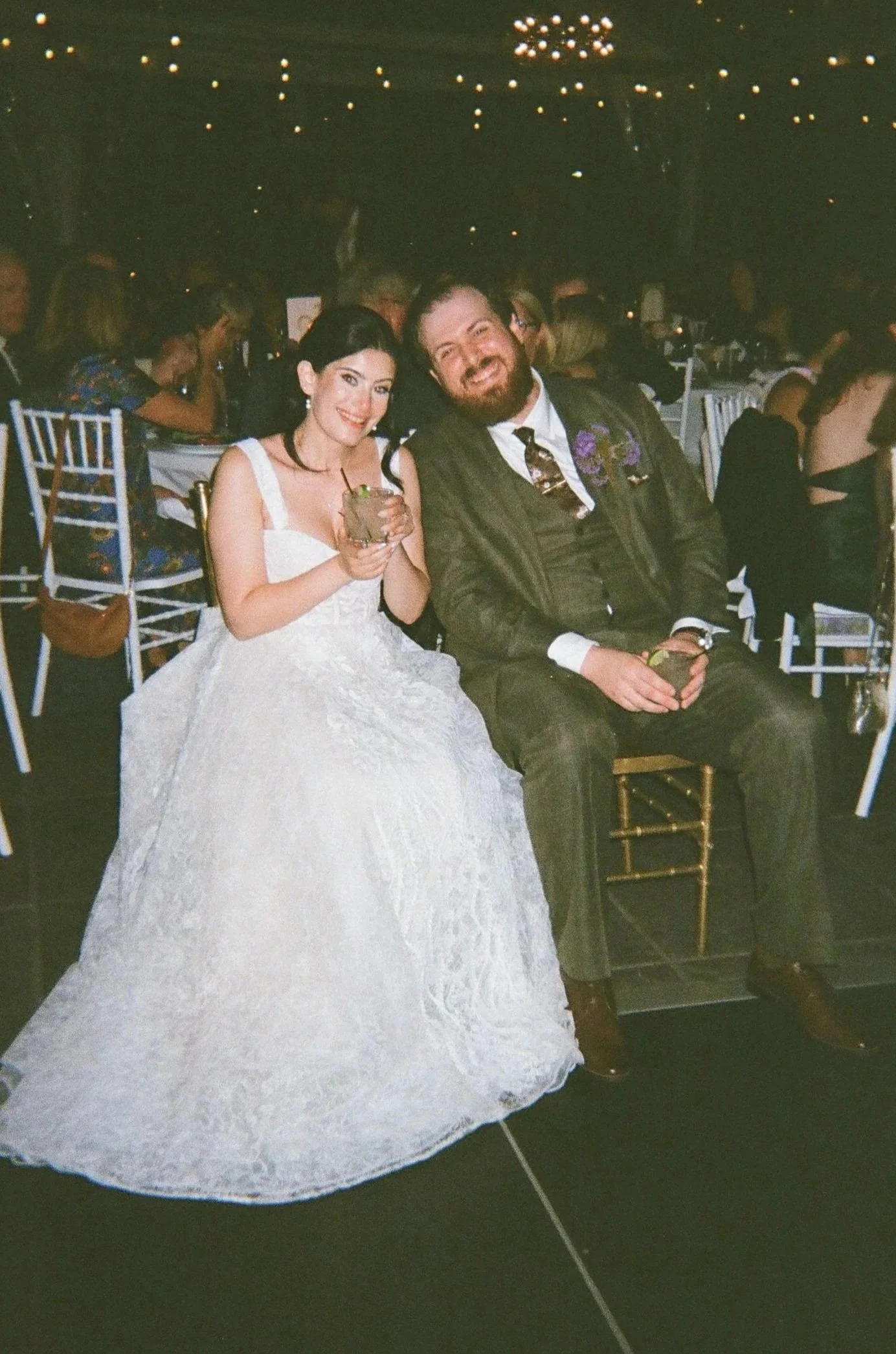 A bride in a white wedding dress and a groom in a dark suit sit together at a wedding reception, smiling and holding drinks, with other guests seated at tables in the background.