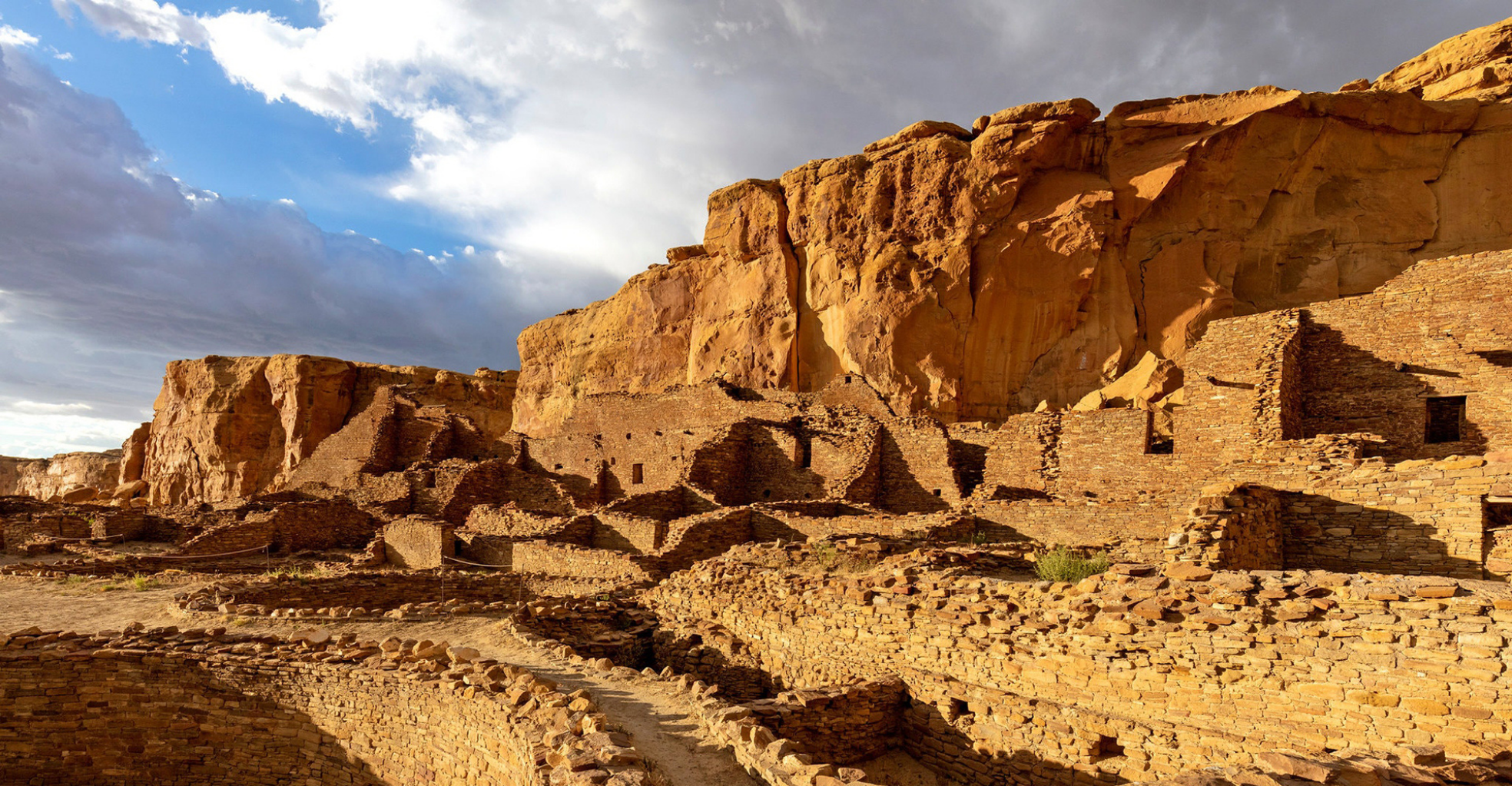 Ancient cliff dwellings built into a sandstone cliff in a desert landscape, with a partly cloudy sky overhead.
