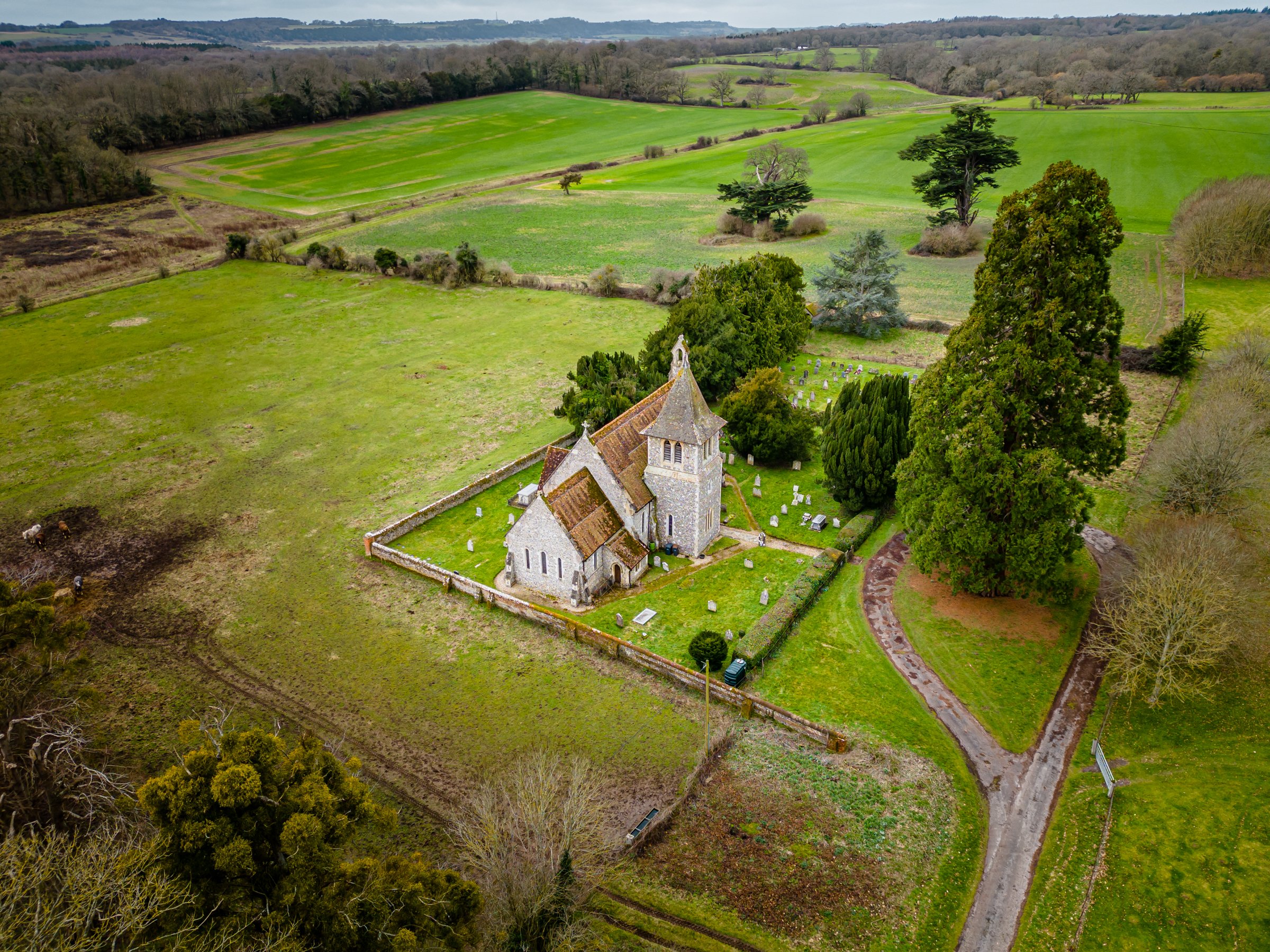  Aerial drone photograph of the 13th century parish church of St Peter’s, East Tytherley, Hampshire.  Showing front elevation view and roof line with partial view of the graveyard. Surrounded by rural landscape of fields. 