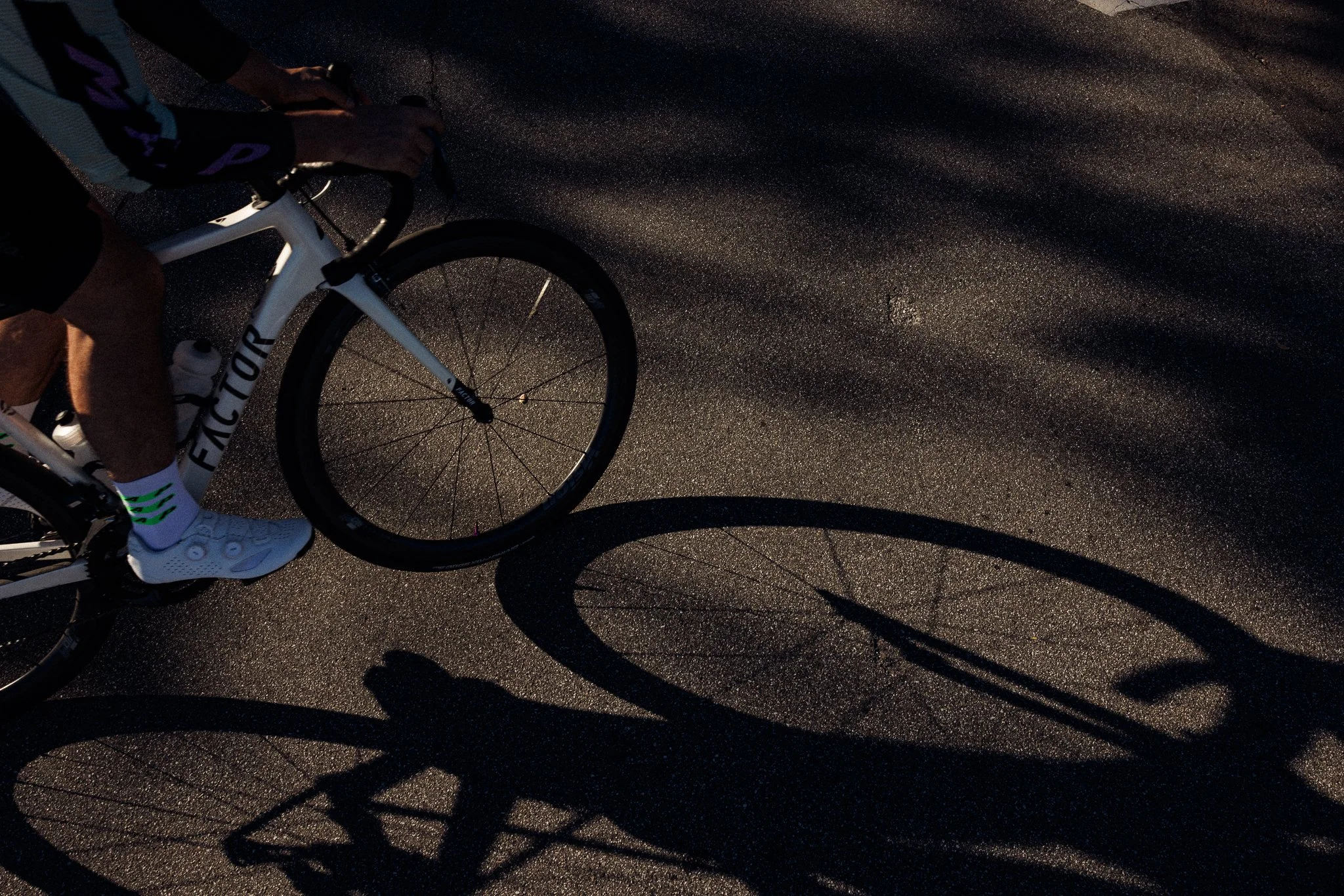 Close-up of a cyclist's legs and white bike, with shadows cast on an asphalt road during low light
