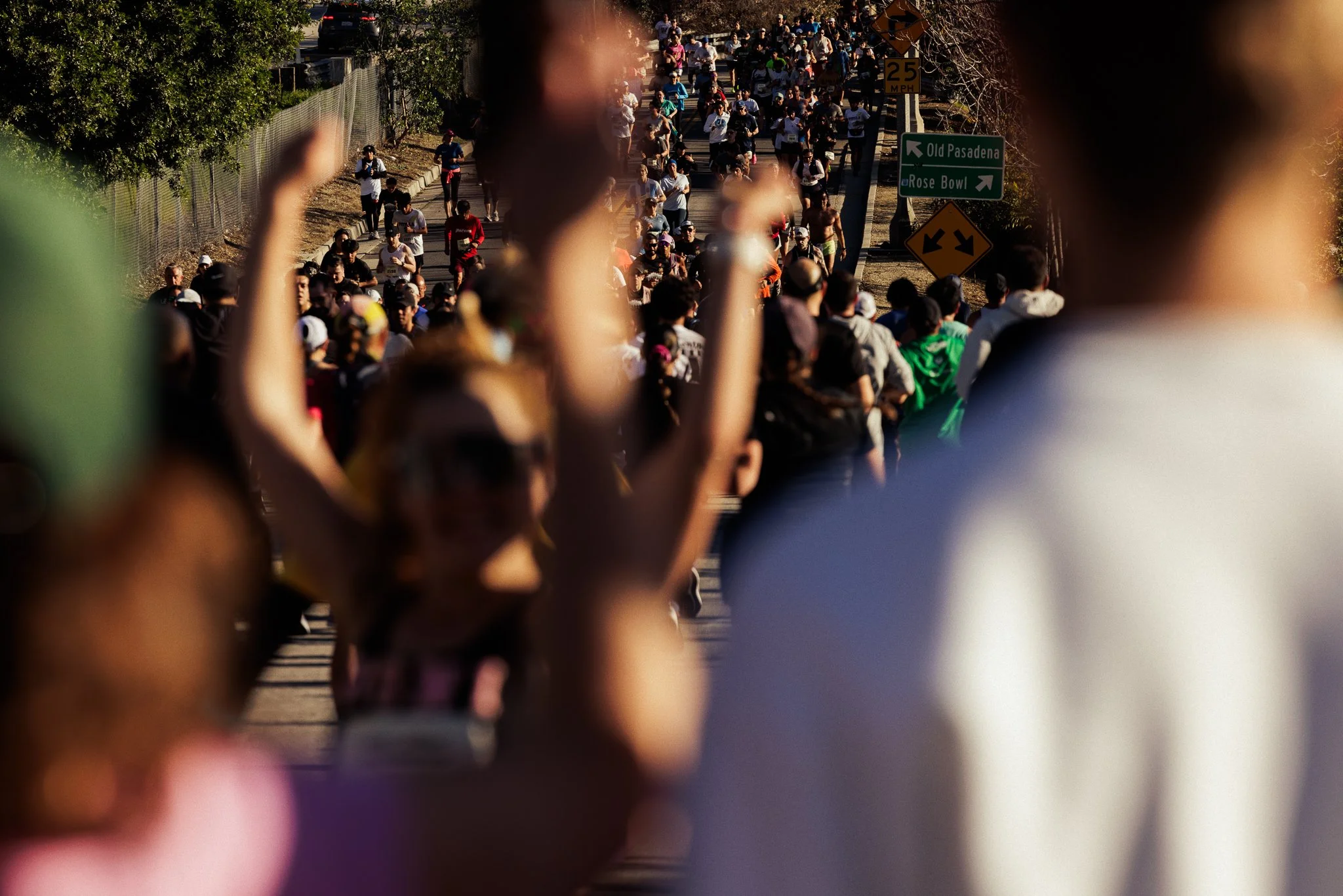 A large group of people walking on a street on a sunny day, with some raising their arms or taking photos, and traffic signs indicating directions to Old Pasadena and Rose Bowl.