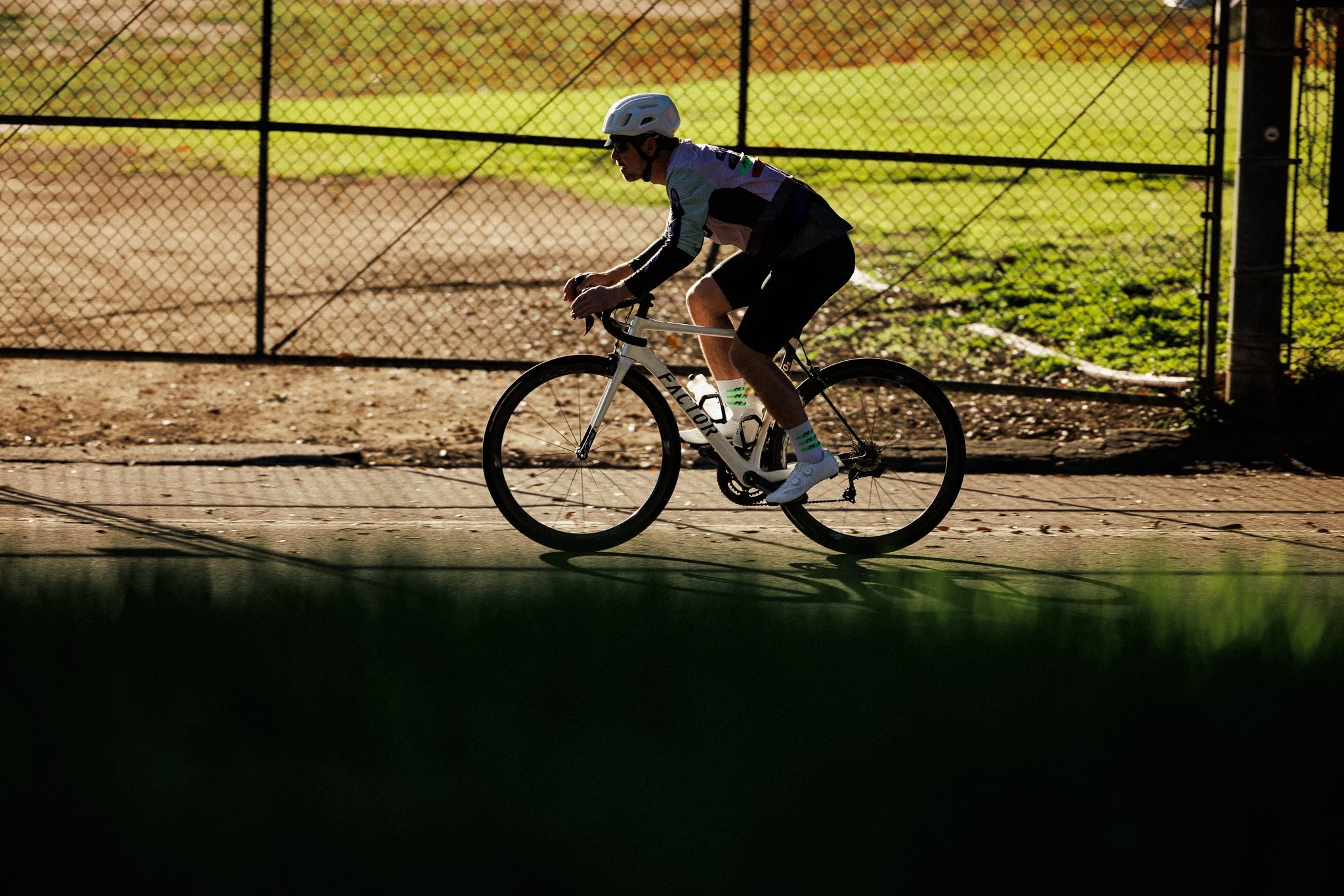A person riding a bicycle on a paved path, wearing a helmet, sunglasses, and athletic clothing, with a grassy field and a chain-link fence in the background.