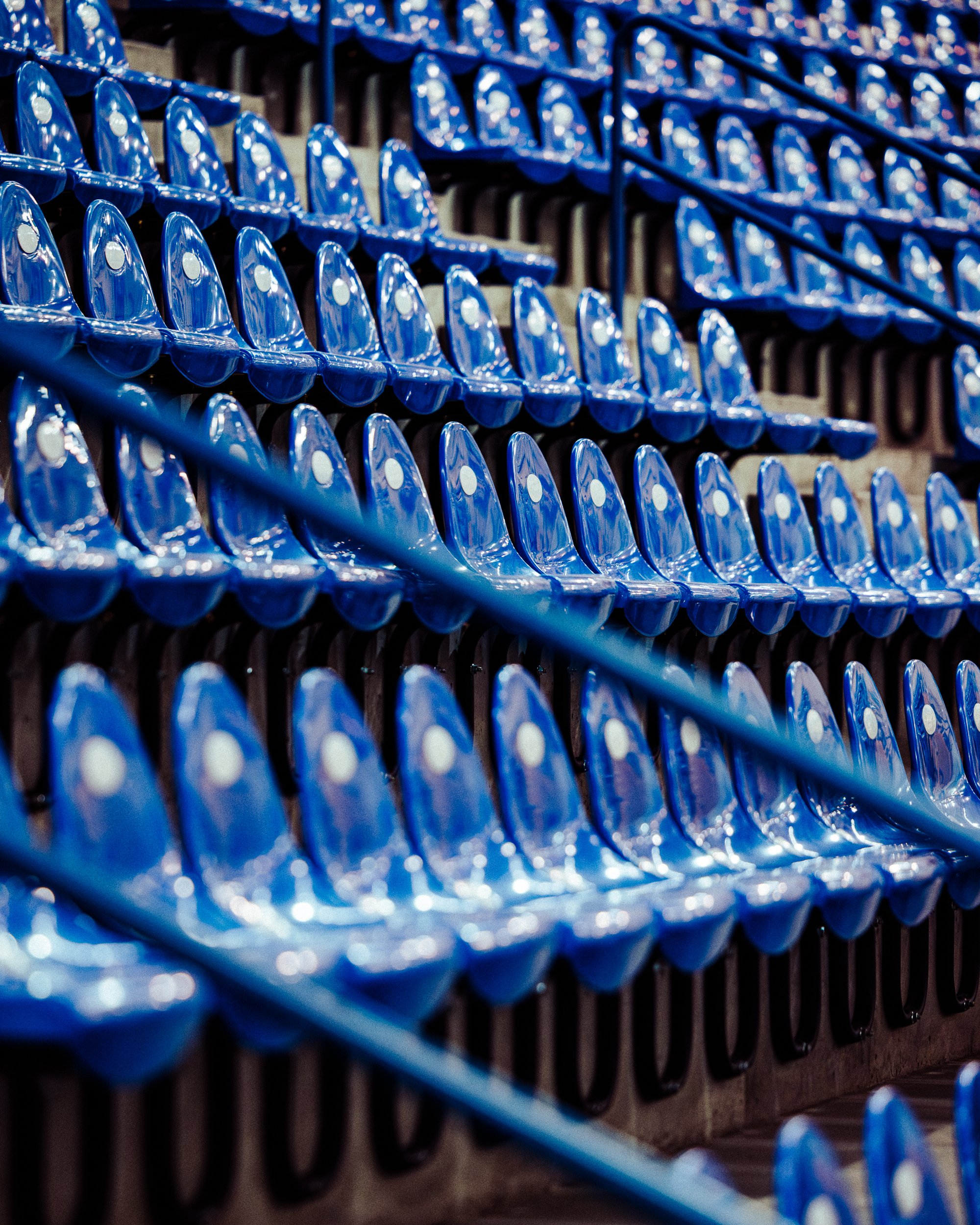 Empty blue stadium seats with black armrests, viewed from an angle with a diagonal blue rail in the foreground.