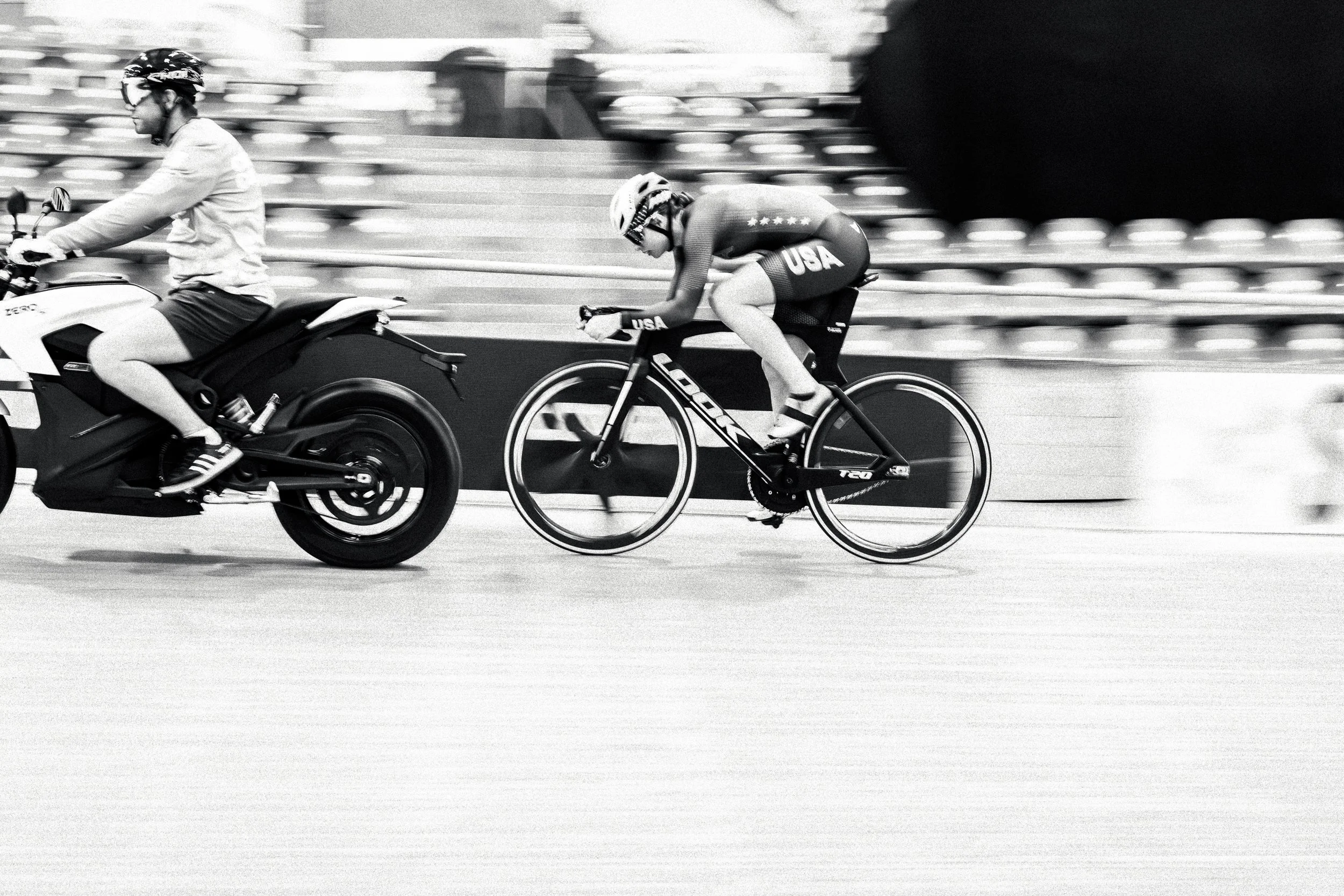 Black and white photo of a cyclist in USA uniform racing on a velodrome track, with a motorbike rider beside him.