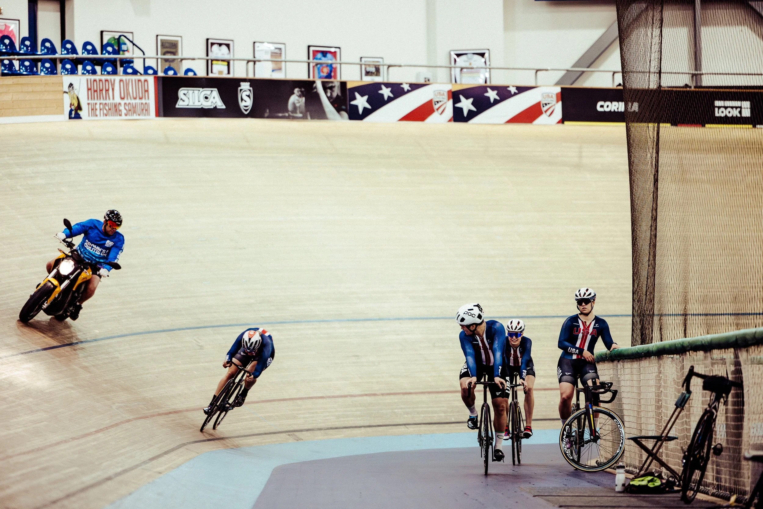 Track cyclists on a velodrome, with some riders racing on the track and others resting against the fence, in a sports venue decorated with banners and flags.