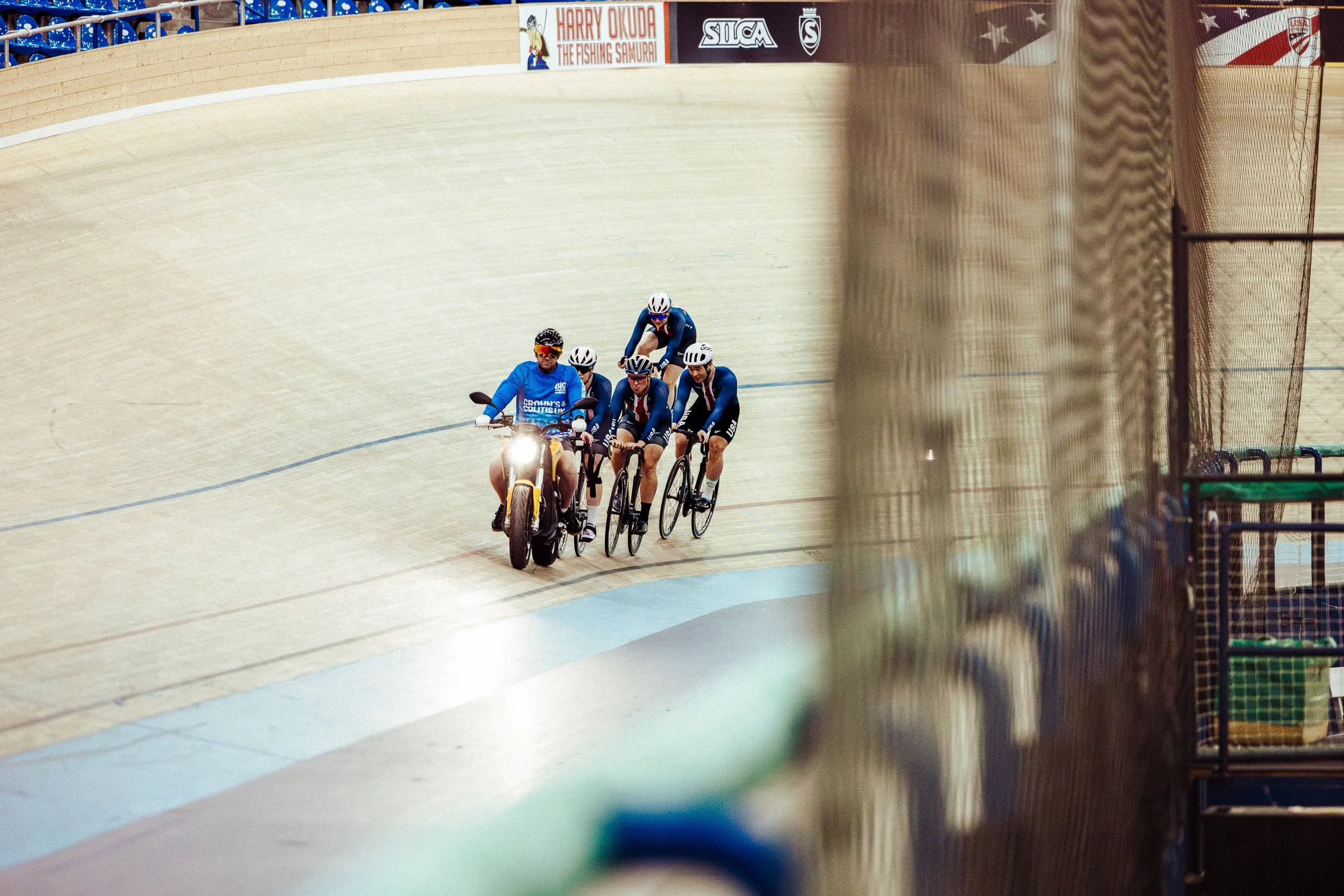 Cyclists and a motorcycle on an indoor velodrome track, with banners and advertisement in the background.