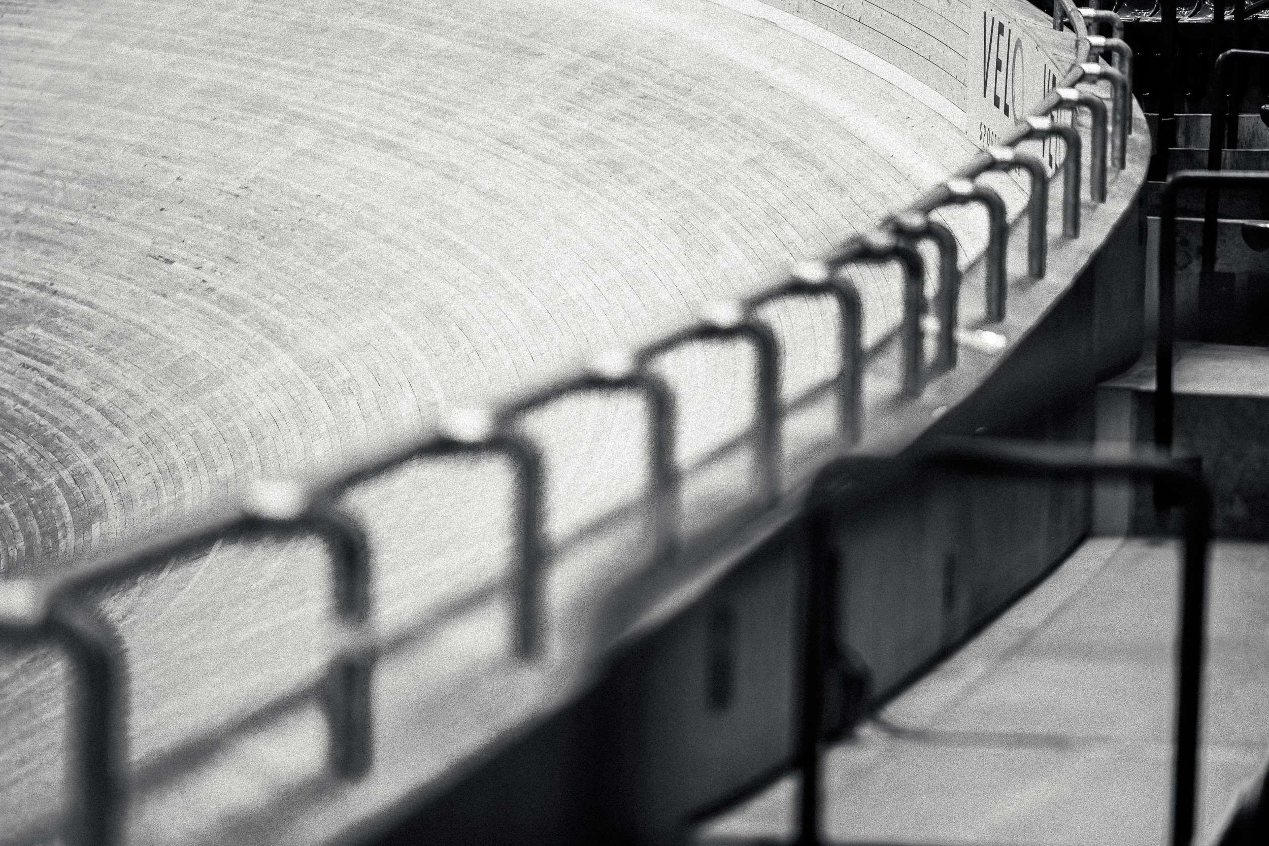 Close-up of a large concrete pipe with metal hooks along the top, in black and white, with some structures and a railing visible in the background.