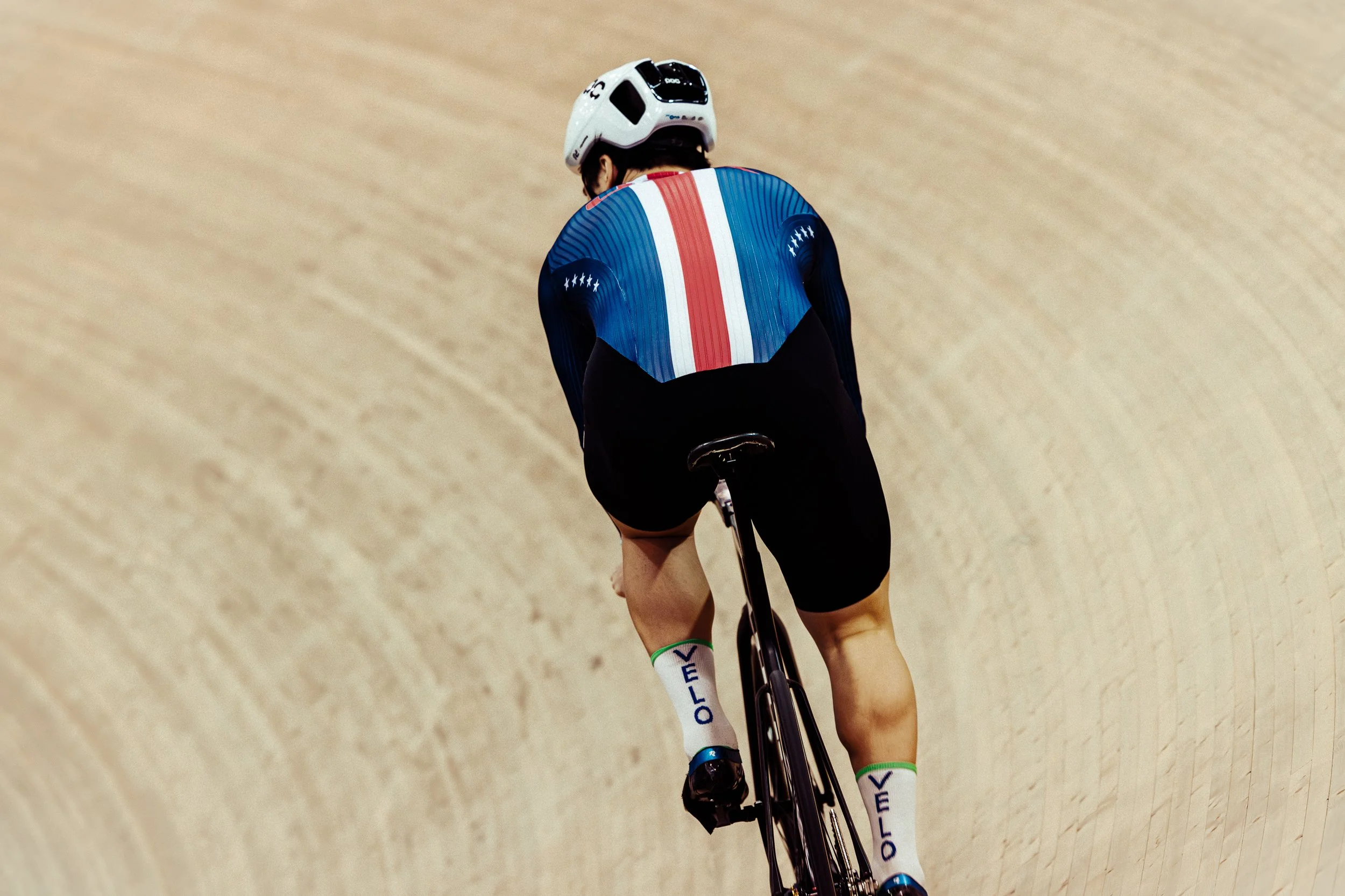 A male cyclist wearing a blue, white, and red jersey and black shorts riding a track bike on a velodrome, viewed from behind.