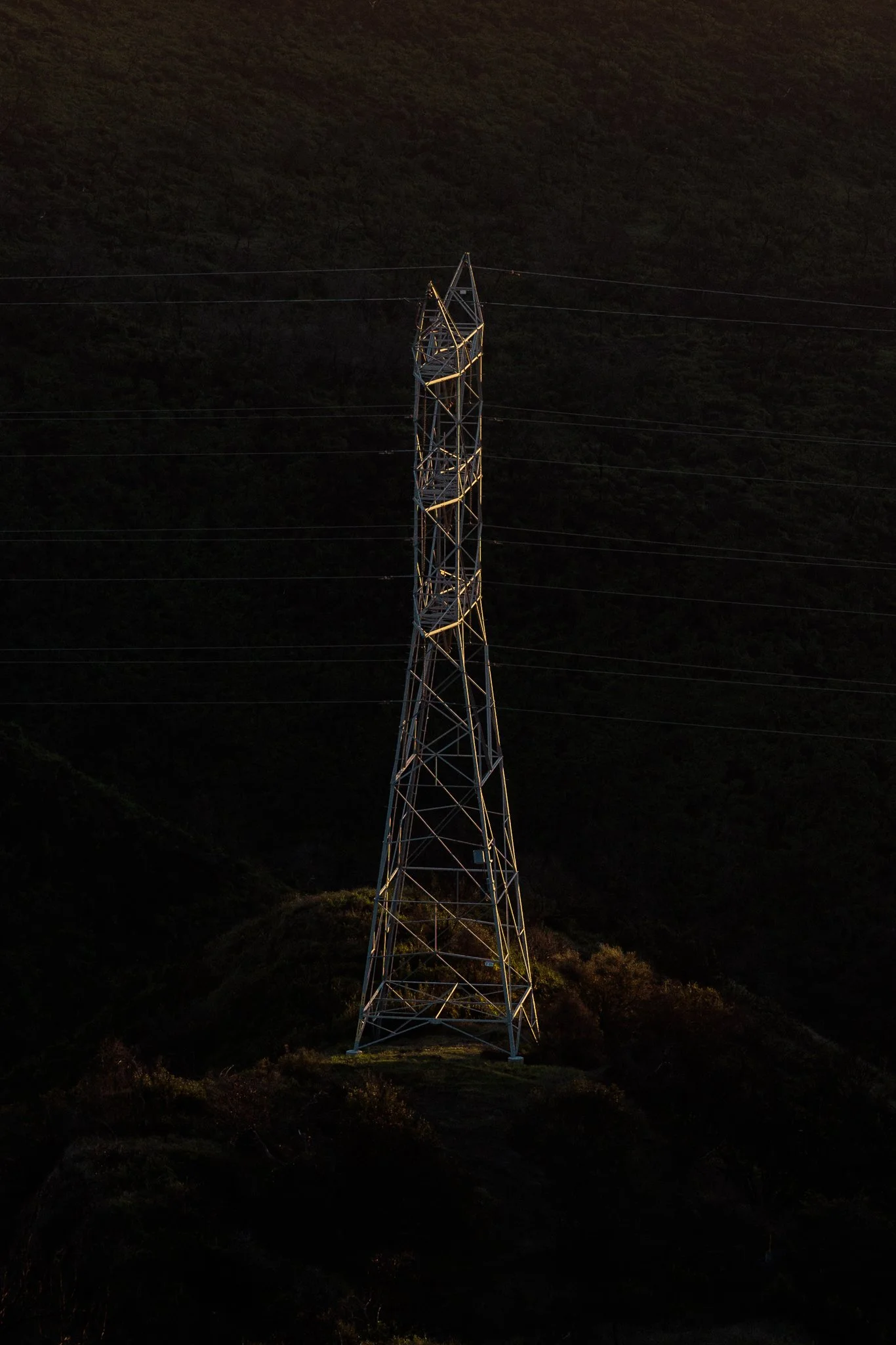 A large metal electrical transmission tower standing on a hillside at dusk, with power lines extending from it.