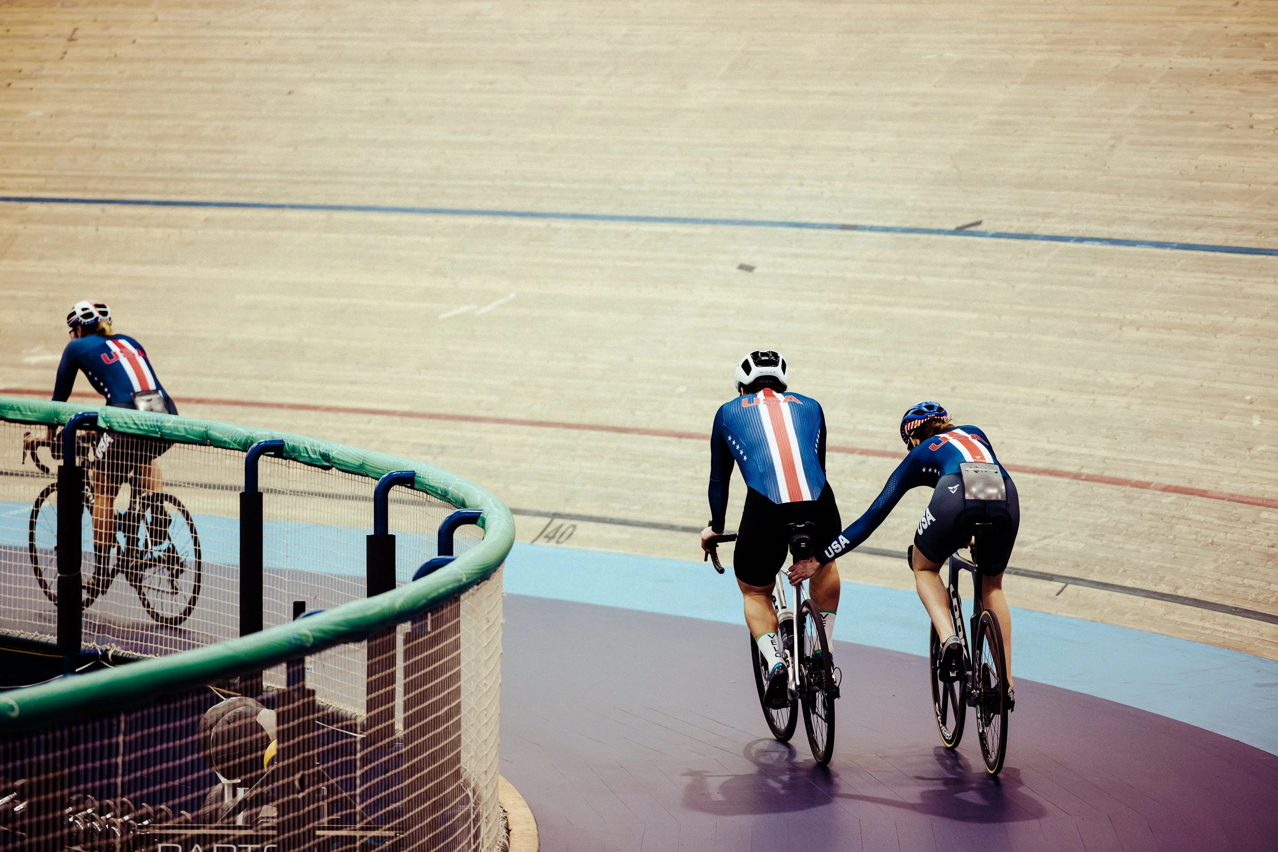 Three cyclists in exchange on a velodrome cycling track, with two of them riding while the third is walking his bike.