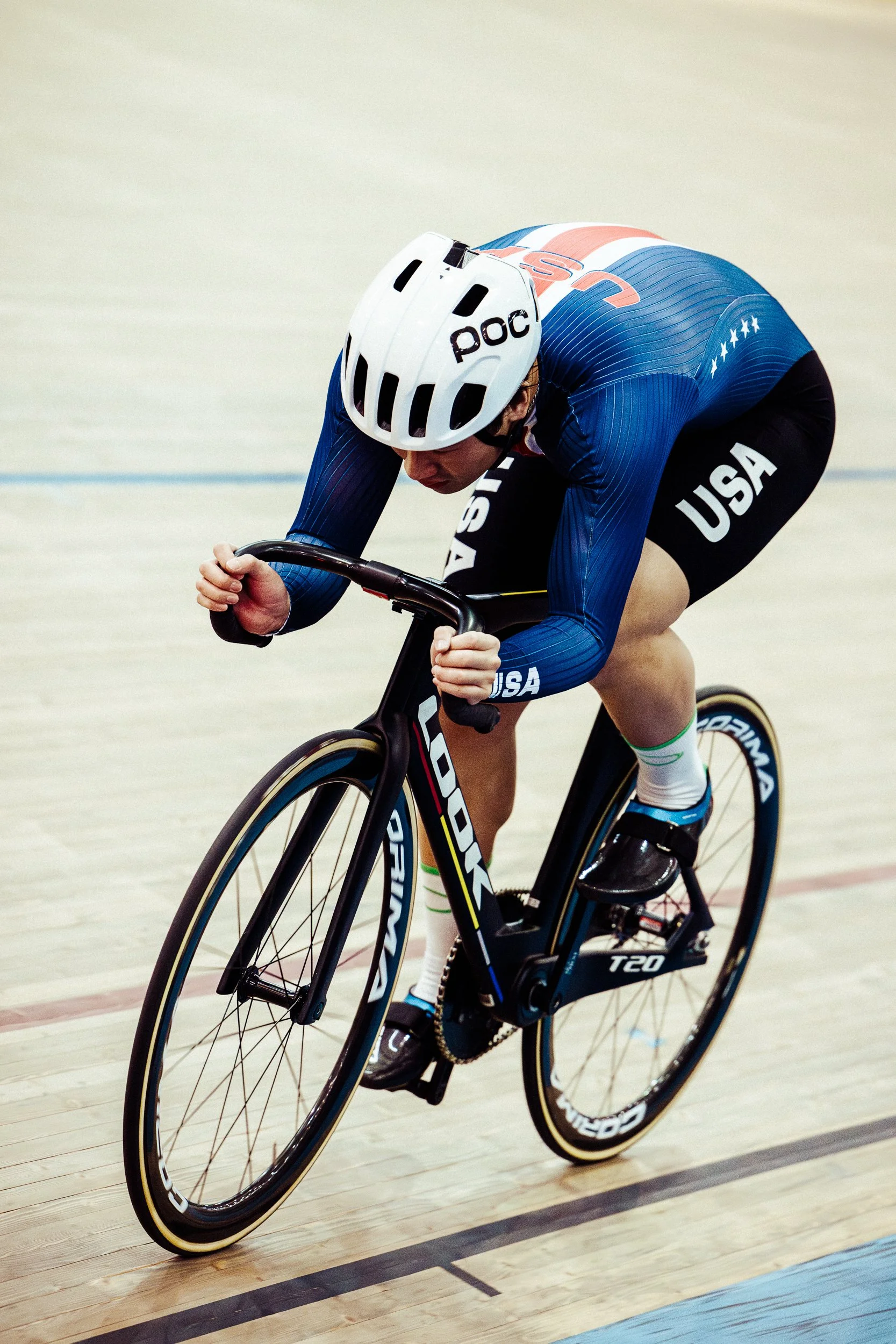 A cyclist wearing a blue USA team uniform and a white helmet is racing on an indoor velodrome cycling track.