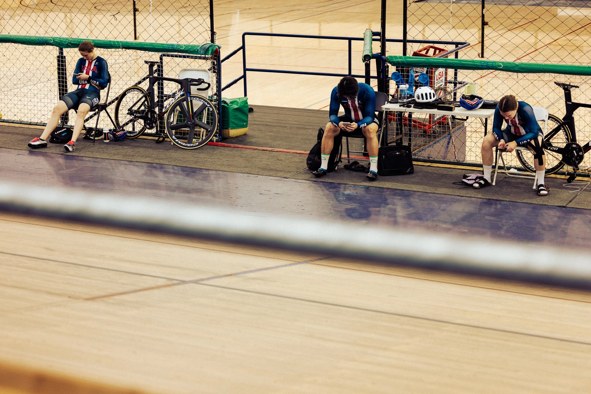 Three male cyclists in Team USA gear sitting on bench or chair at indoor velodrome with bicycles parked behind them, electronics on table, and bike helmet on table, with a netted fence in the background.