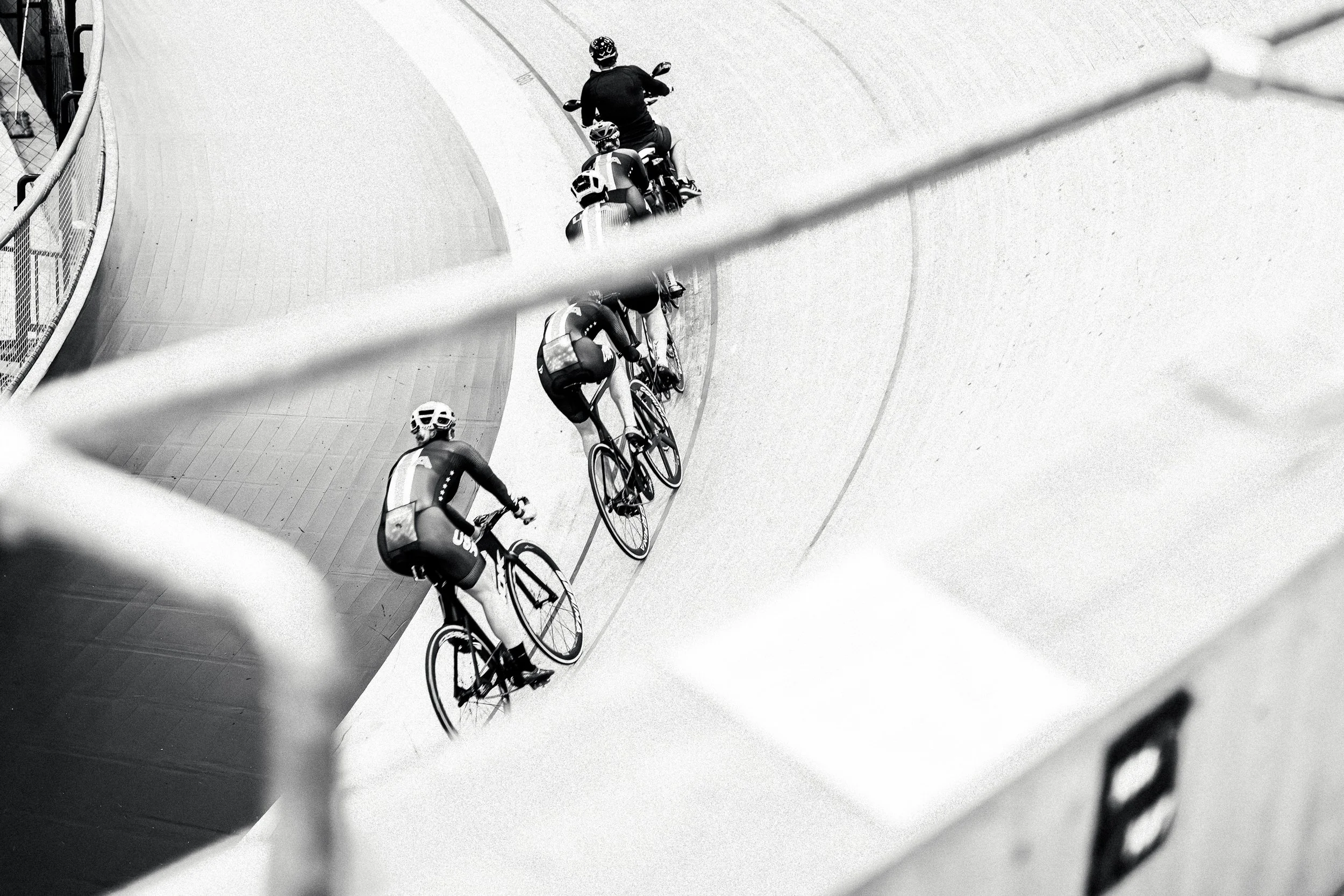 Black and white photo of four cyclists riding on a velodrome track, viewed through a fence.