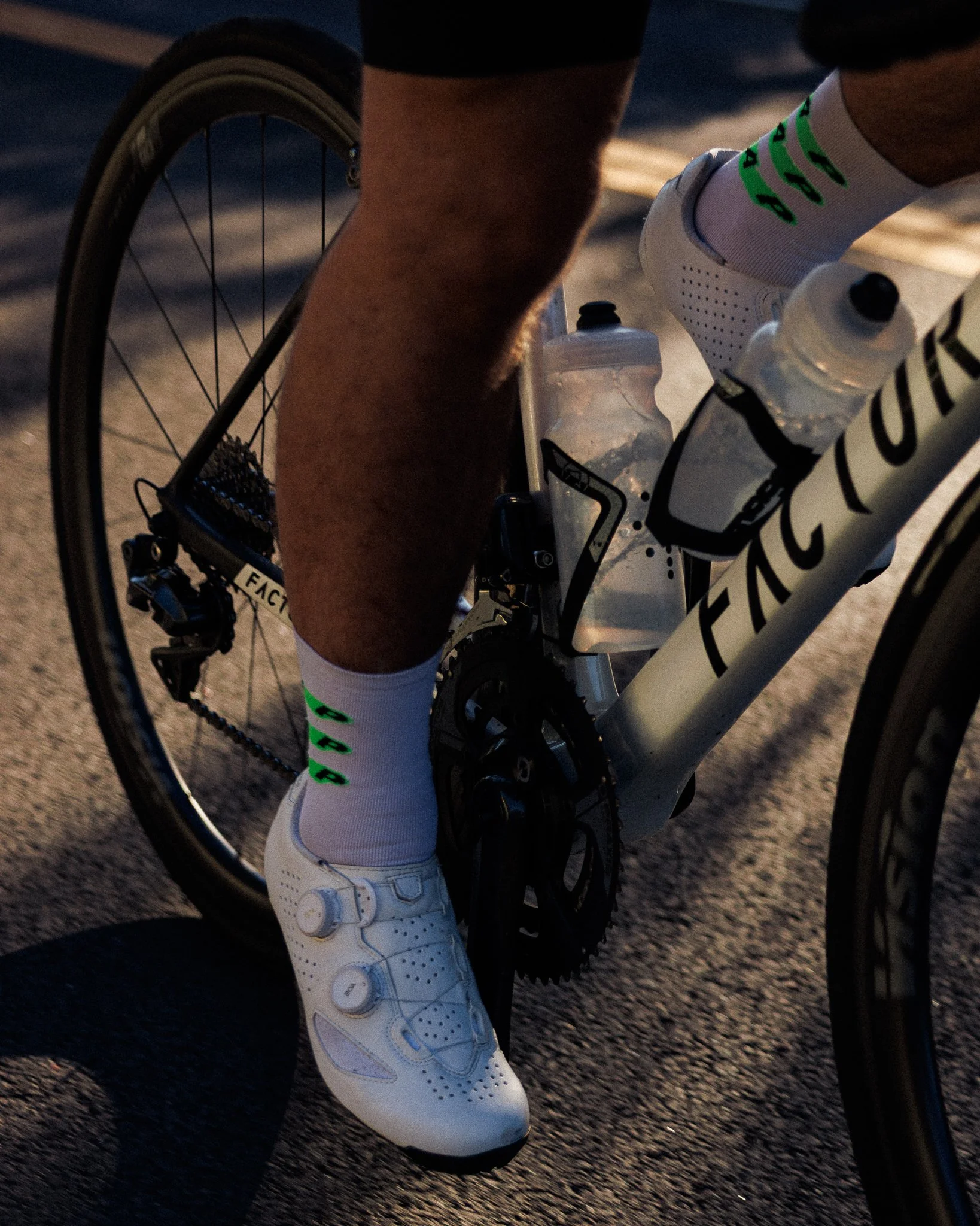 Close-up of a cyclist's leg, white cycling shoe clipped into pedal, white socks with green logo, on a road bike with water bottles on frame, during dusk or dawn.