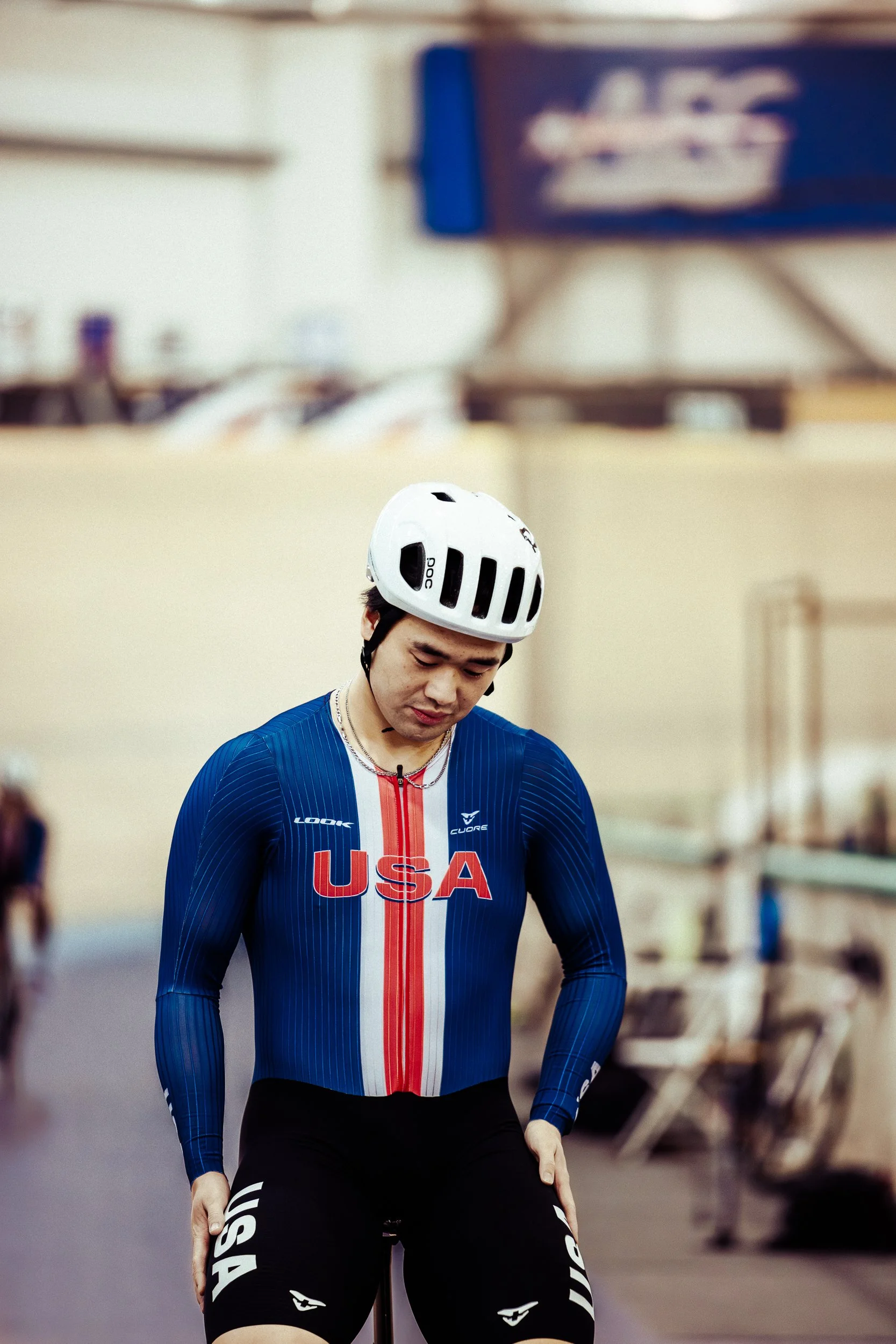 A male athlete wearing a blue USA cycling jersey with red and white stripes, black shorts with white USA logos, and a white helmet, is looking down while preparing to ride a bike indoors.