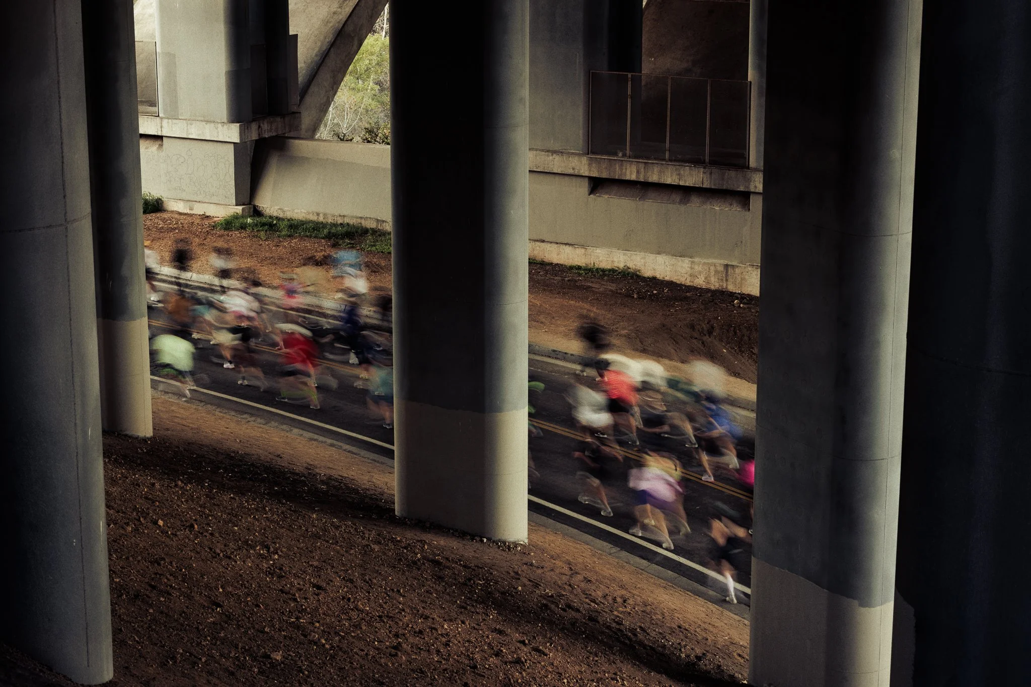 A group of cyclists riding on a road beneath a bridge with large concrete pillars.