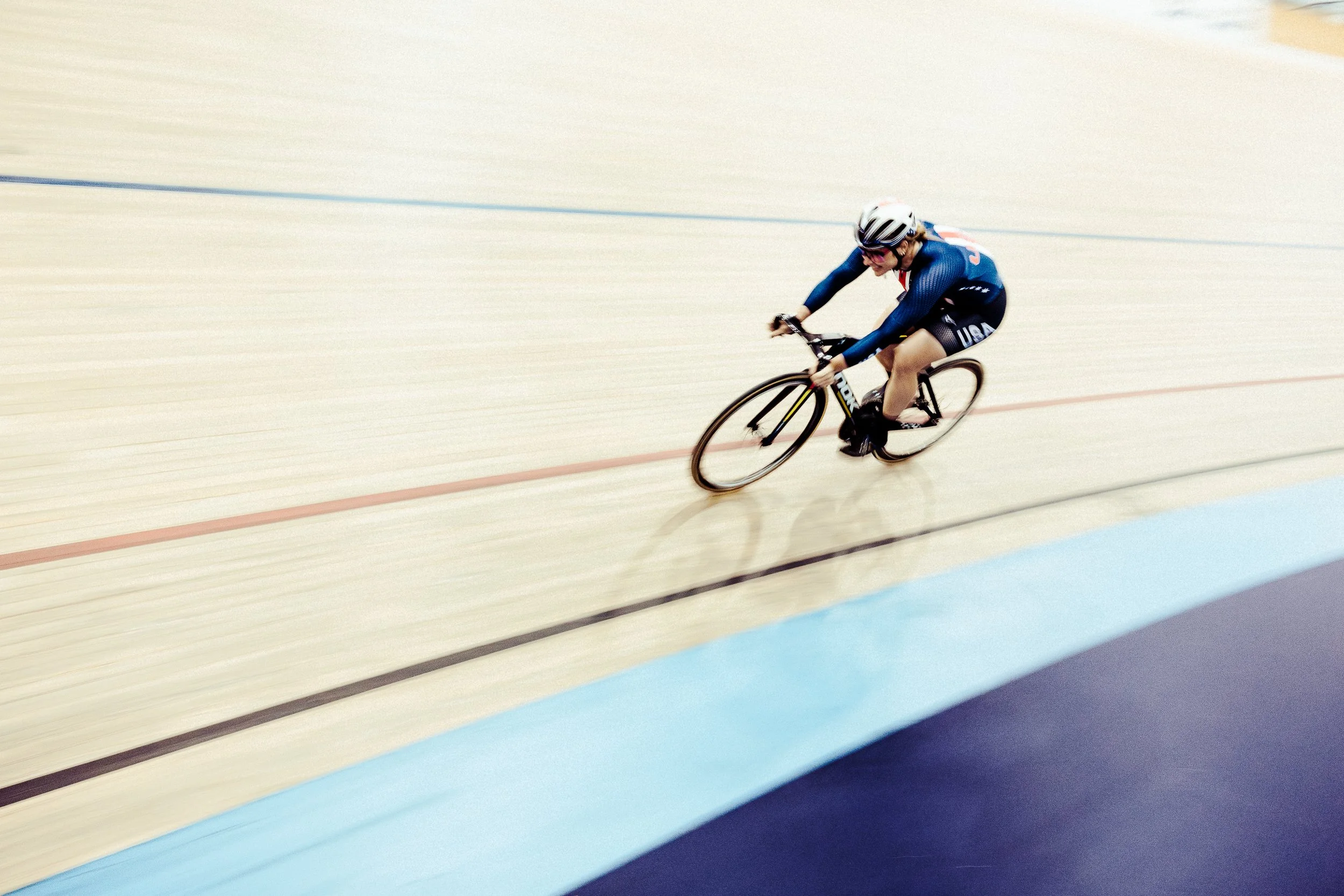 A female cyclist wearing a blue USA jersey and helmet racing on an indoor velodrome track.