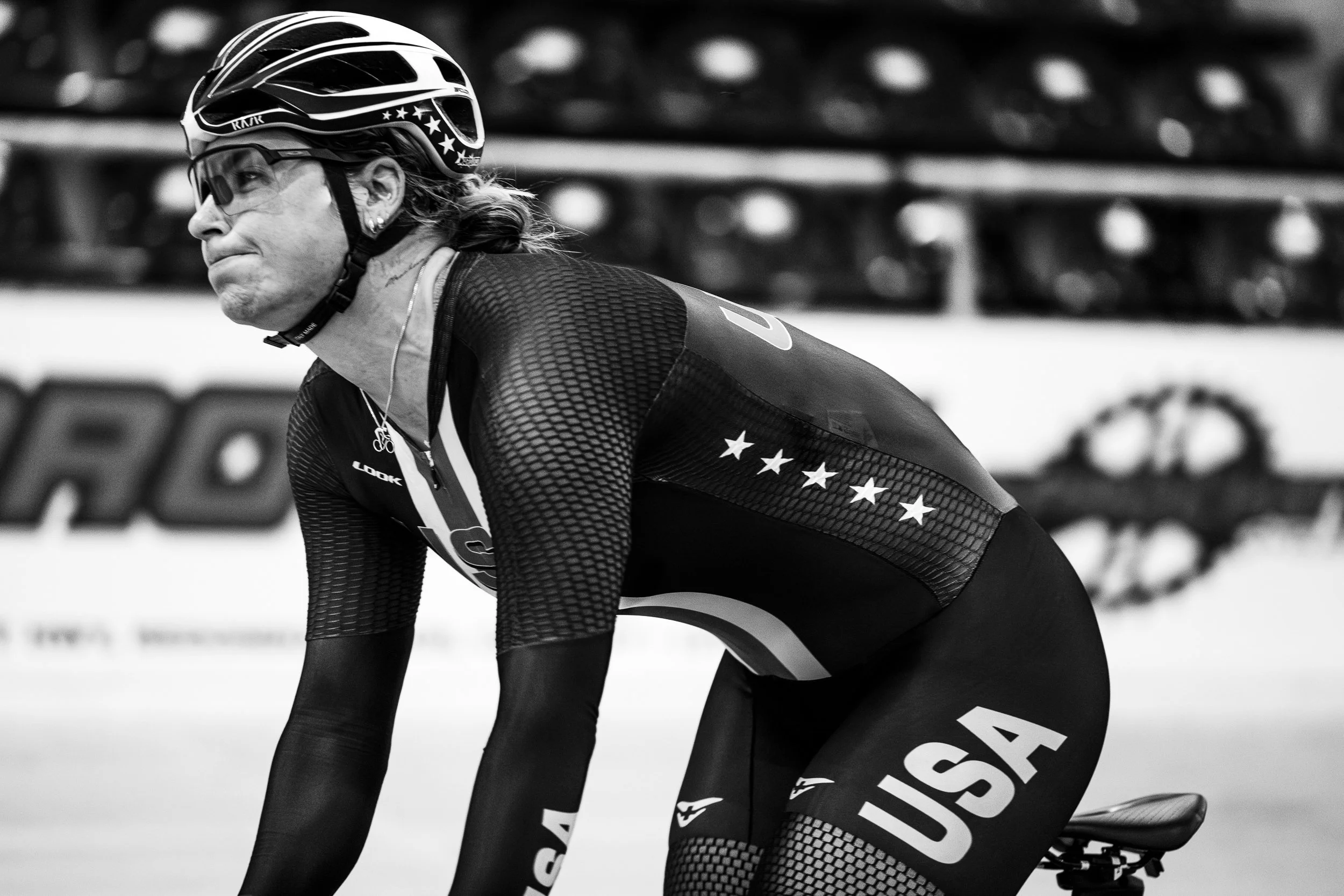 A female cyclist wearing sunglasses, a helmet, and a USA team jersey, crouching on a bike during a race or training session.