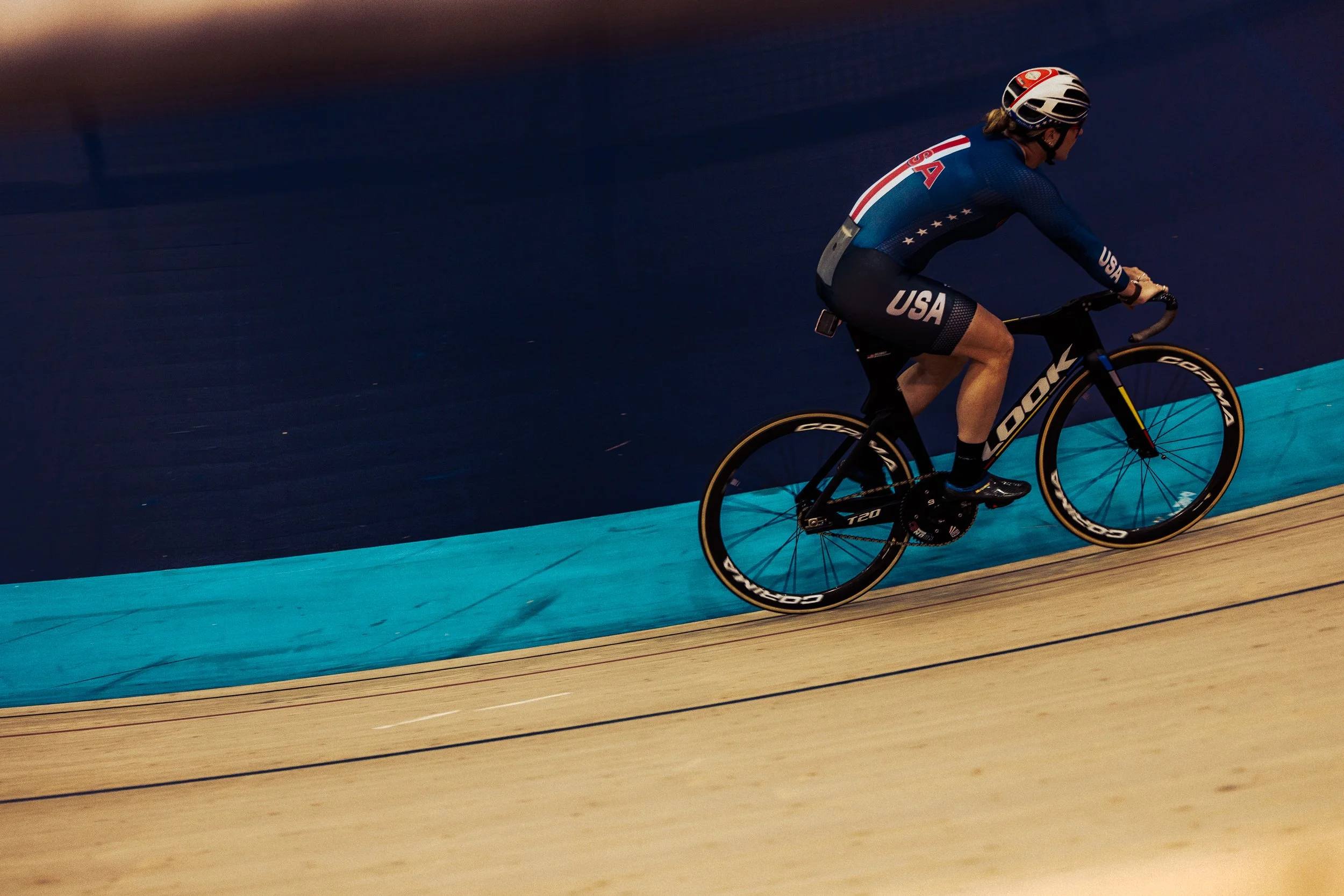 A cyclist wearing a USA team uniform riding a black track bike on an indoor velodrome with a blue and tan track.