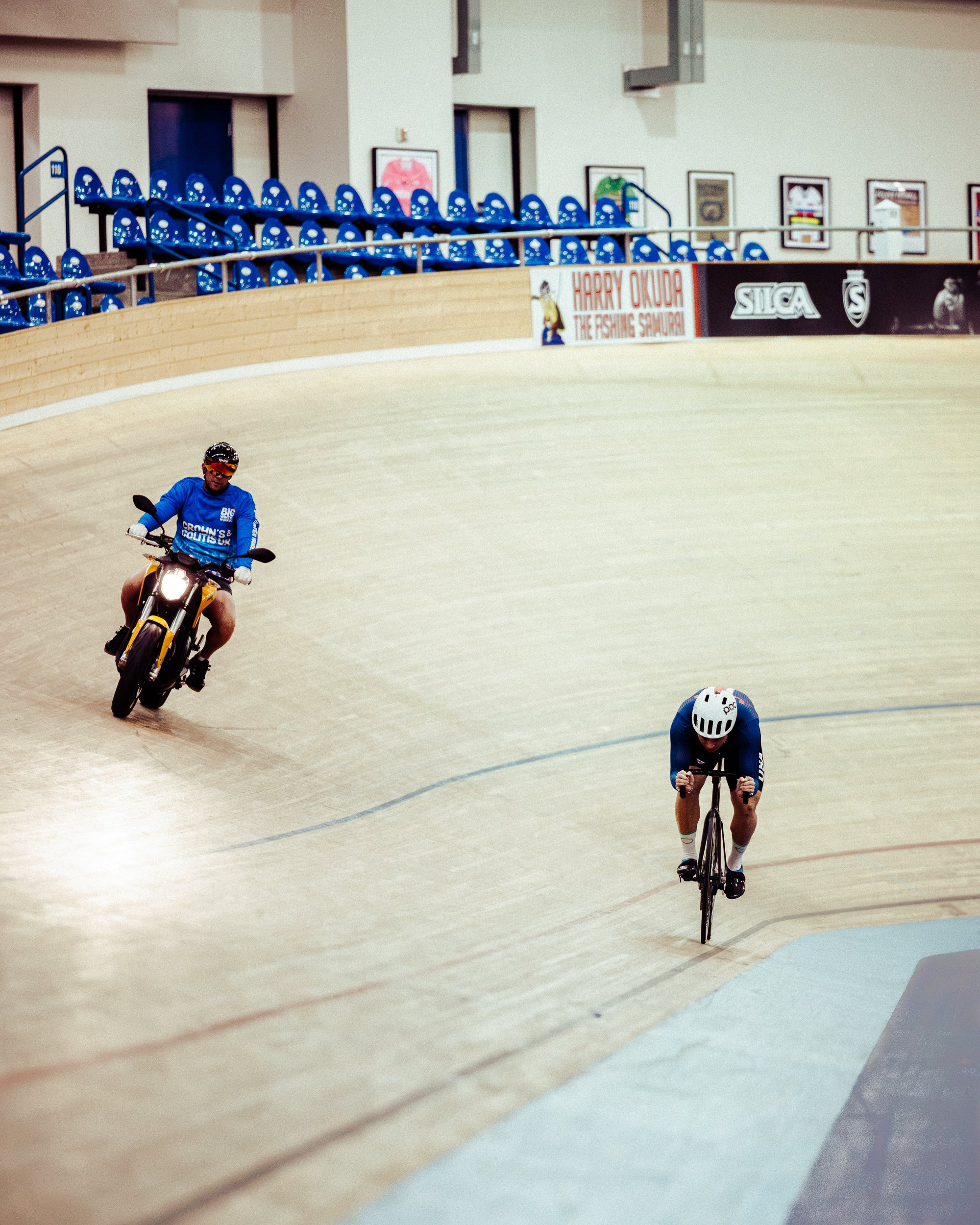 An indoor velodrome with two cyclists and a motorcycle. One cyclist is leaning forward on their bike, wearing a helmet and blue athletic gear. The motorcycle rider is following behind, wearing a helmet and sunglasses, also dressed in blue.