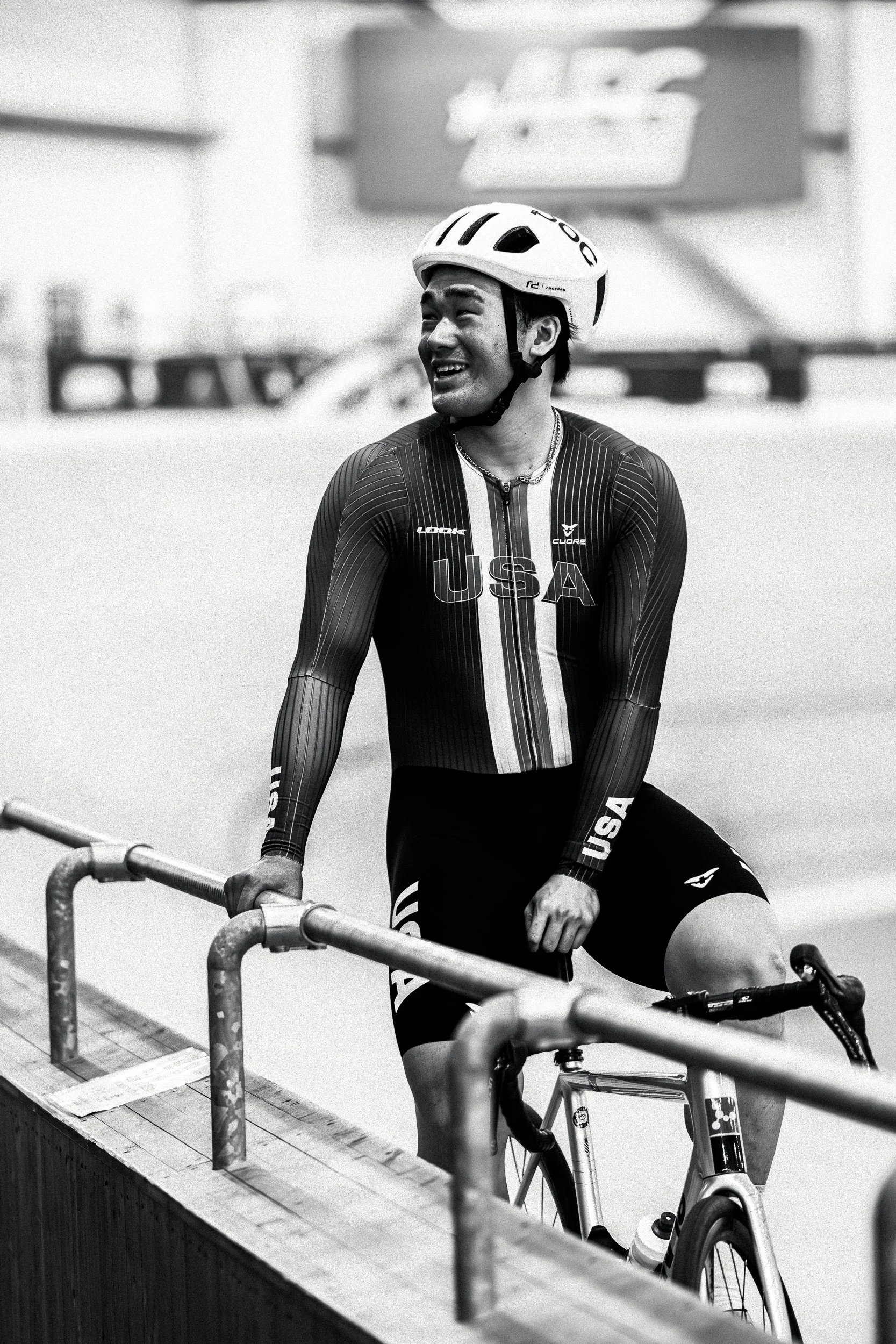 A male cyclist wearing a helmet and USA sportswear, smiling while holding his bicycle indoors.