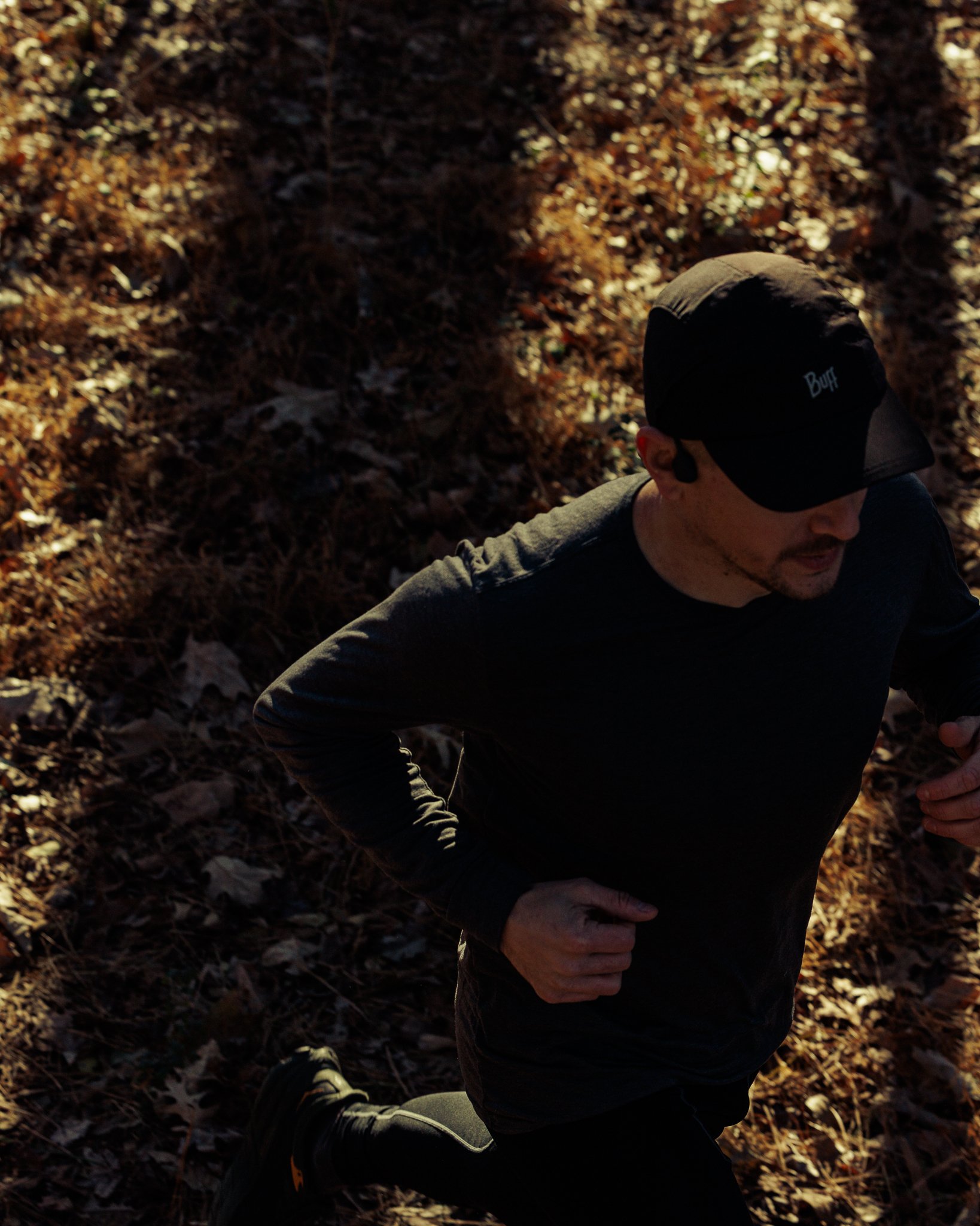 A man running outdoor during sunlight filtering through trees, dressed in dark athletic wear, wearing a black cap and sports earbuds, on a leaf-covered trail.
