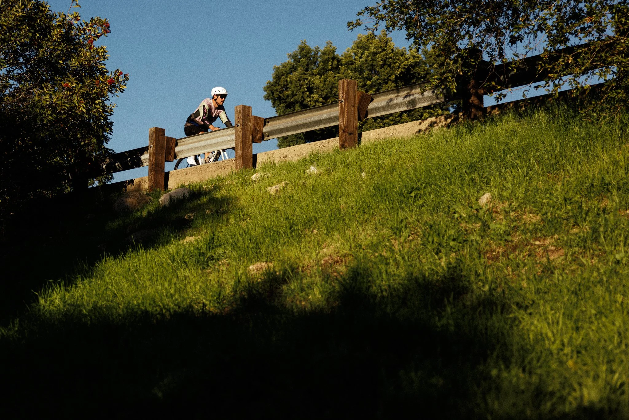 A person riding a bicycle up a grassy hill next to a wooden and metal guardrail on a sunny day with clear blue sky and trees in the background.