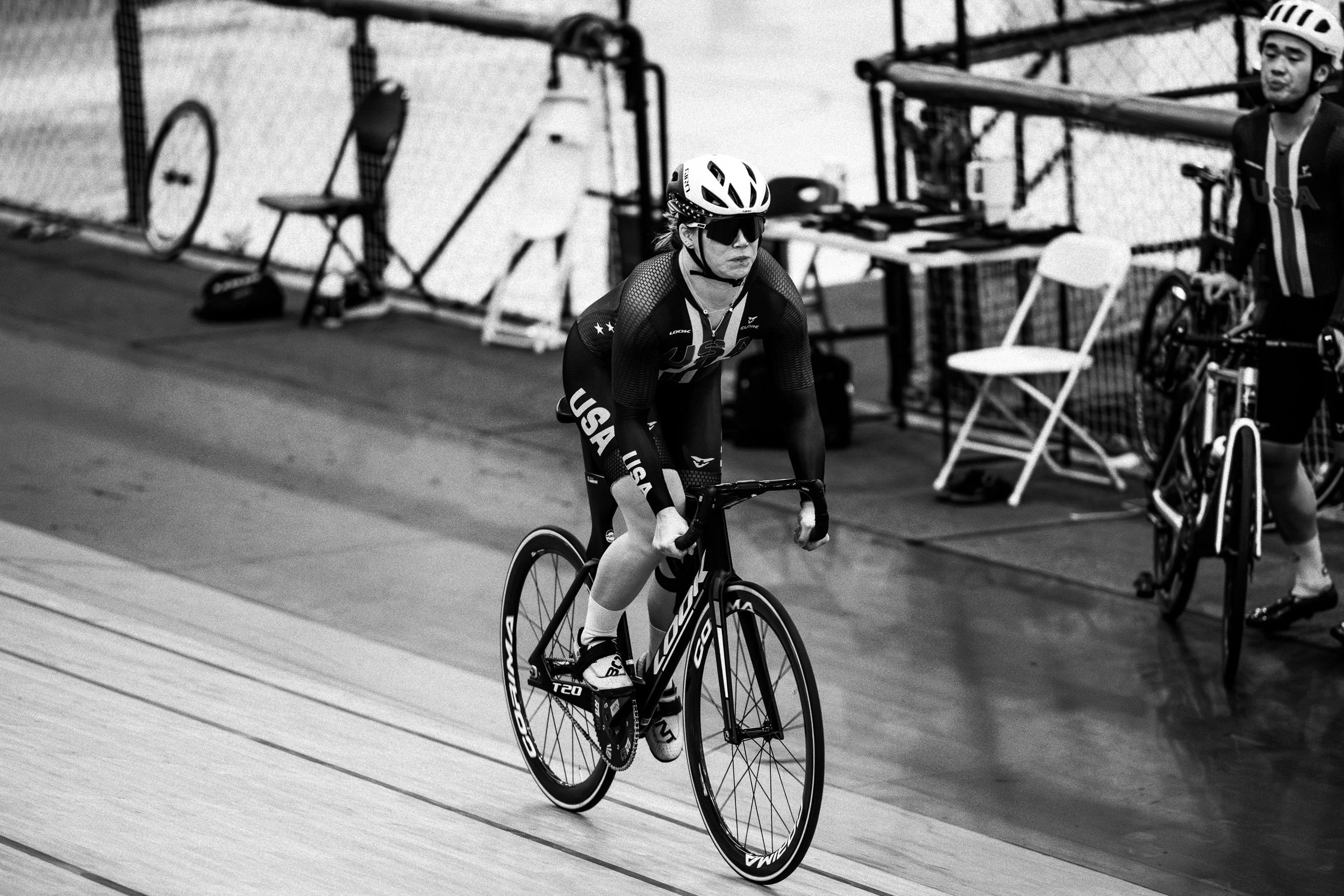 A female cyclist wearing sunglasses and USA team gear riding a black Goka track bicycle on an indoor velodrome track.